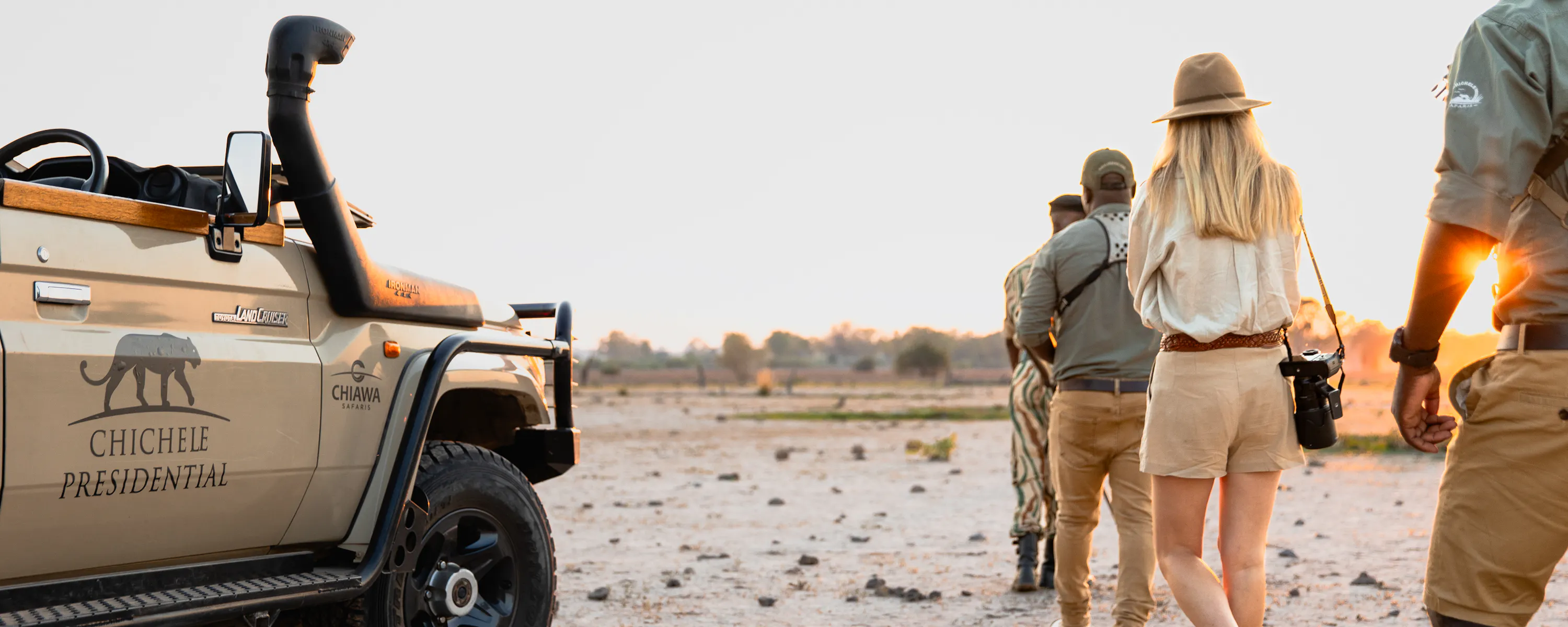 Guided walking safari at sunset with guests and rangers beside a safari vehicle in an open landscape.