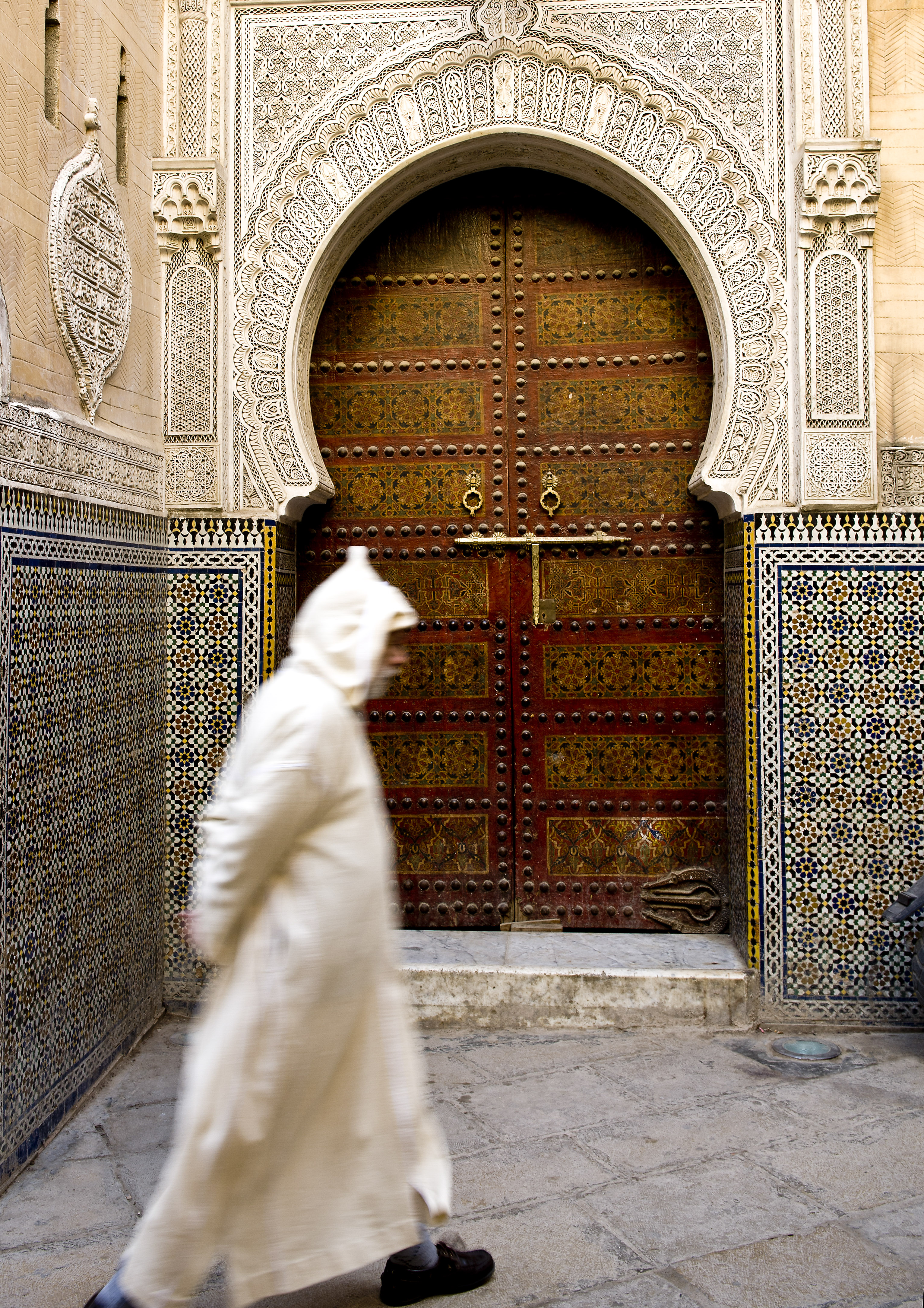 Person walking past an intricately carved doorway with mosaic tilework in Fes.