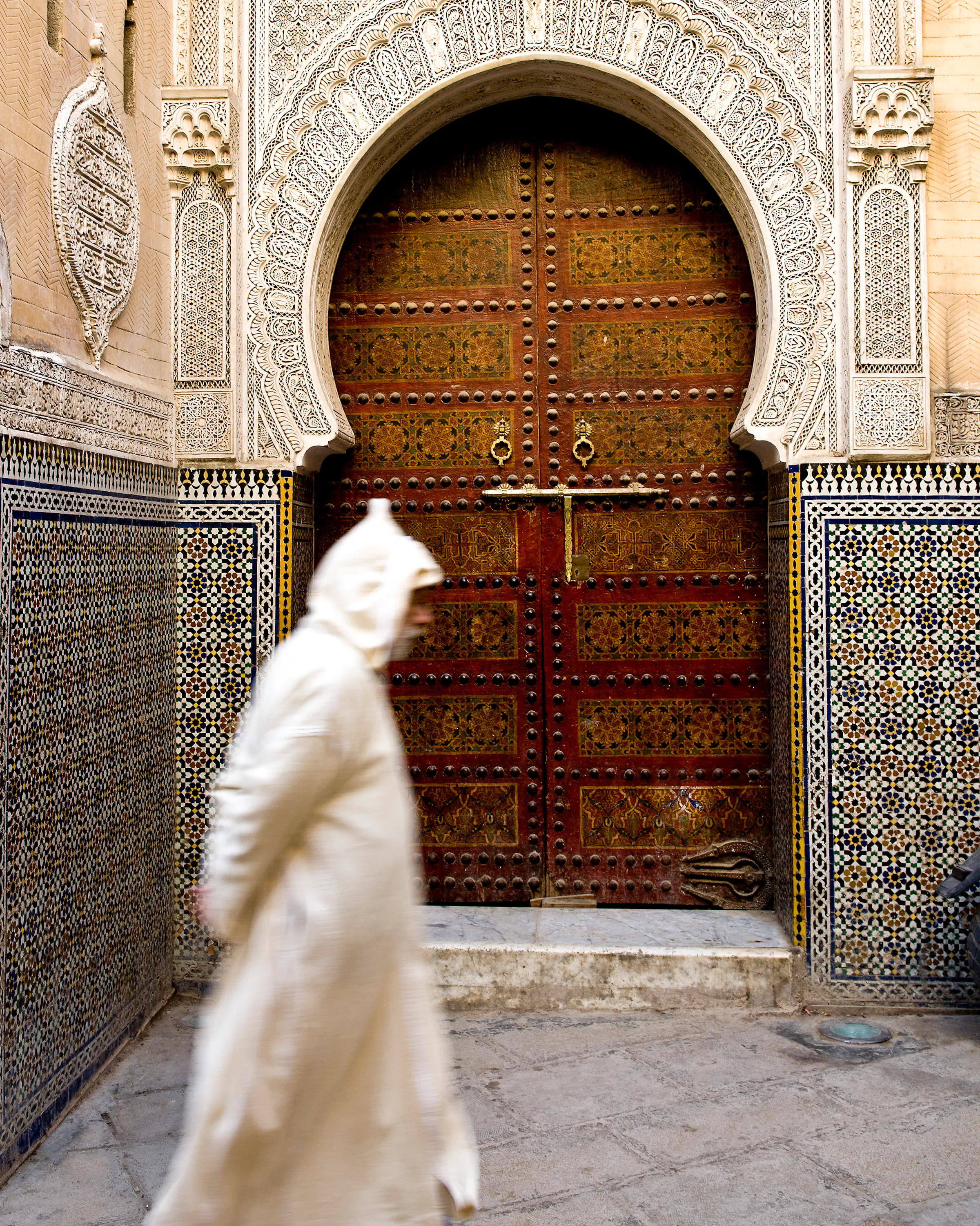 Person walking past an intricately carved doorway with mosaic tilework in Fes.