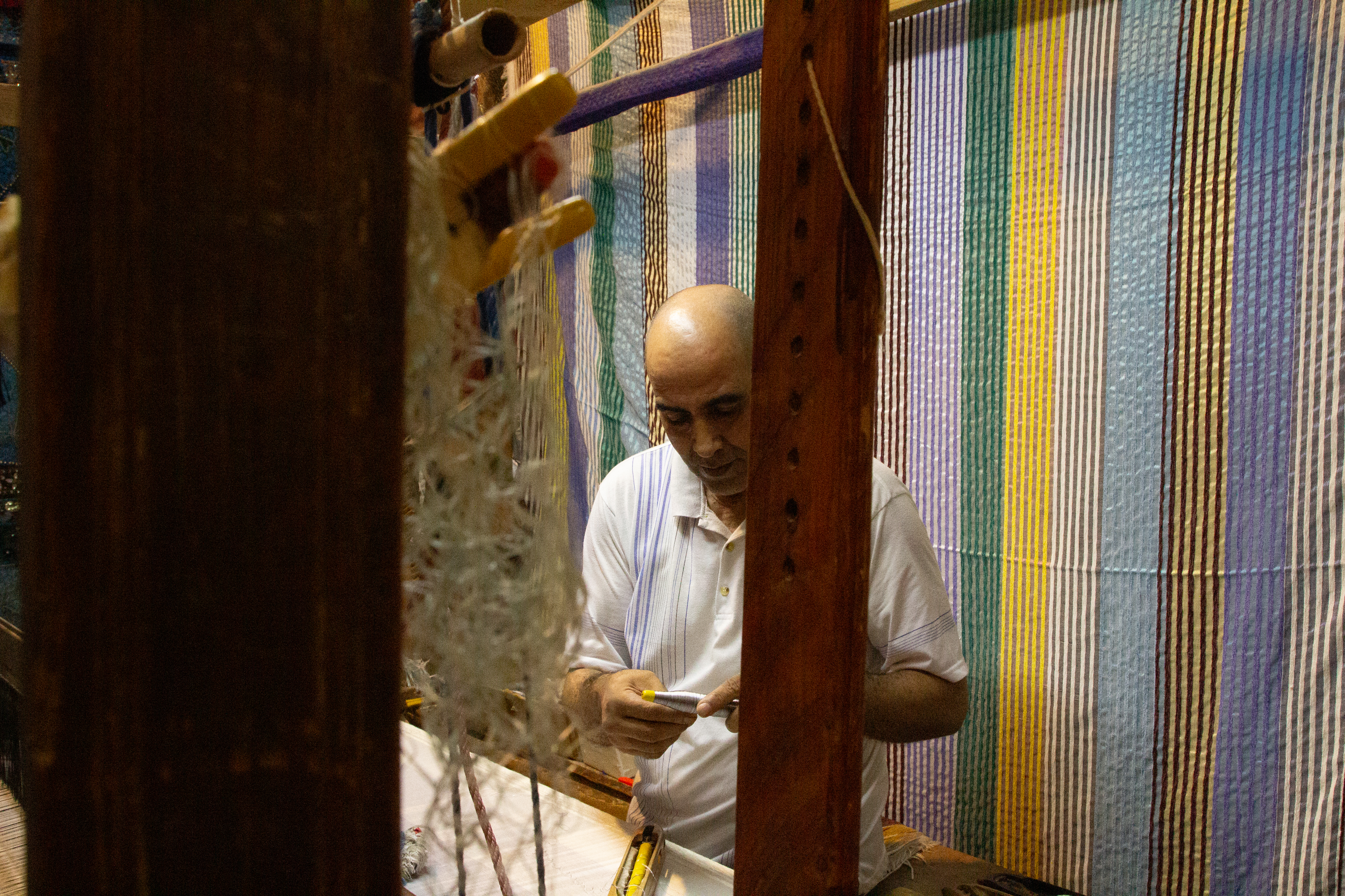 Artisan working at a loom, weaving fabric with striped textiles hanging behind.