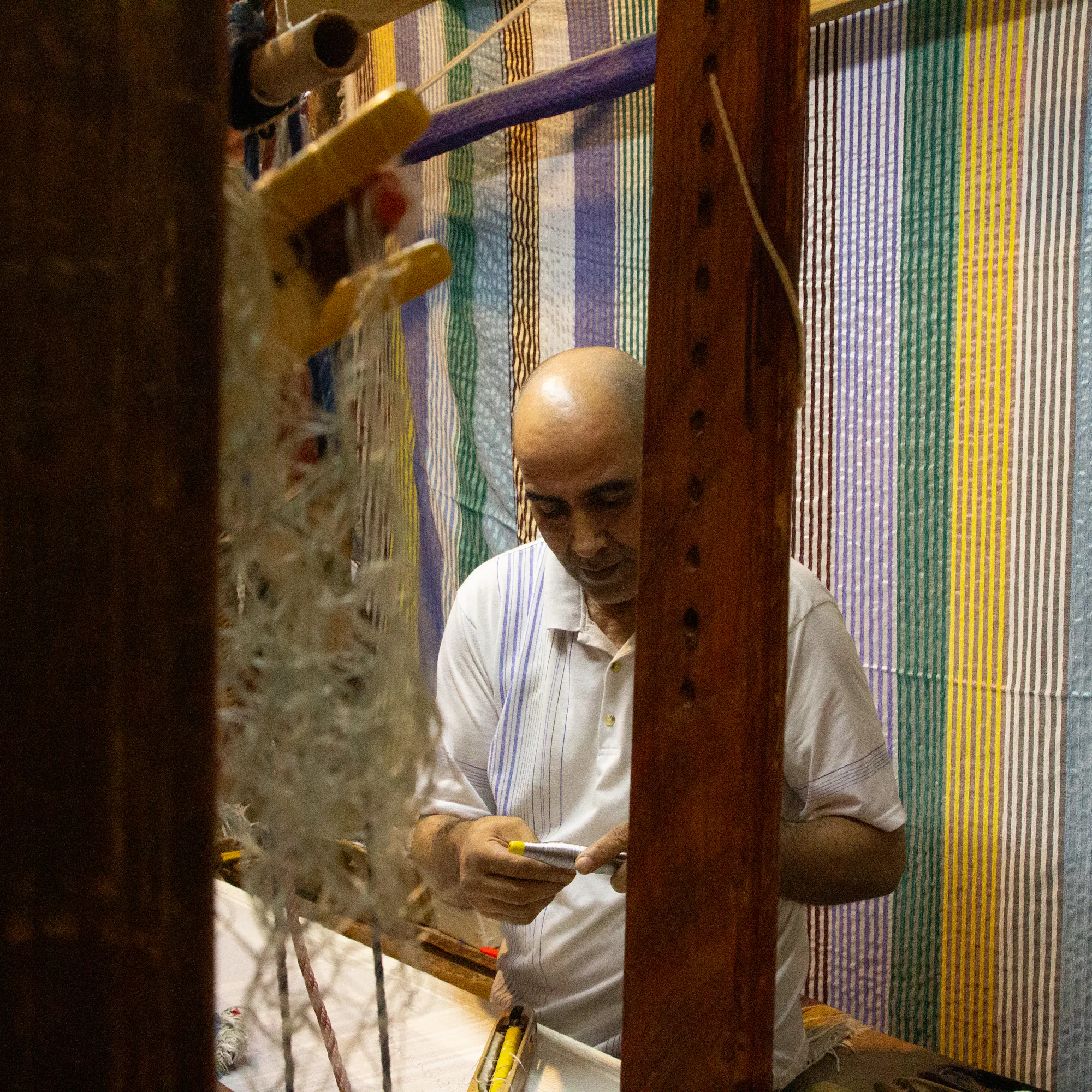 Artisan working at a loom, weaving fabric with striped textiles hanging behind.