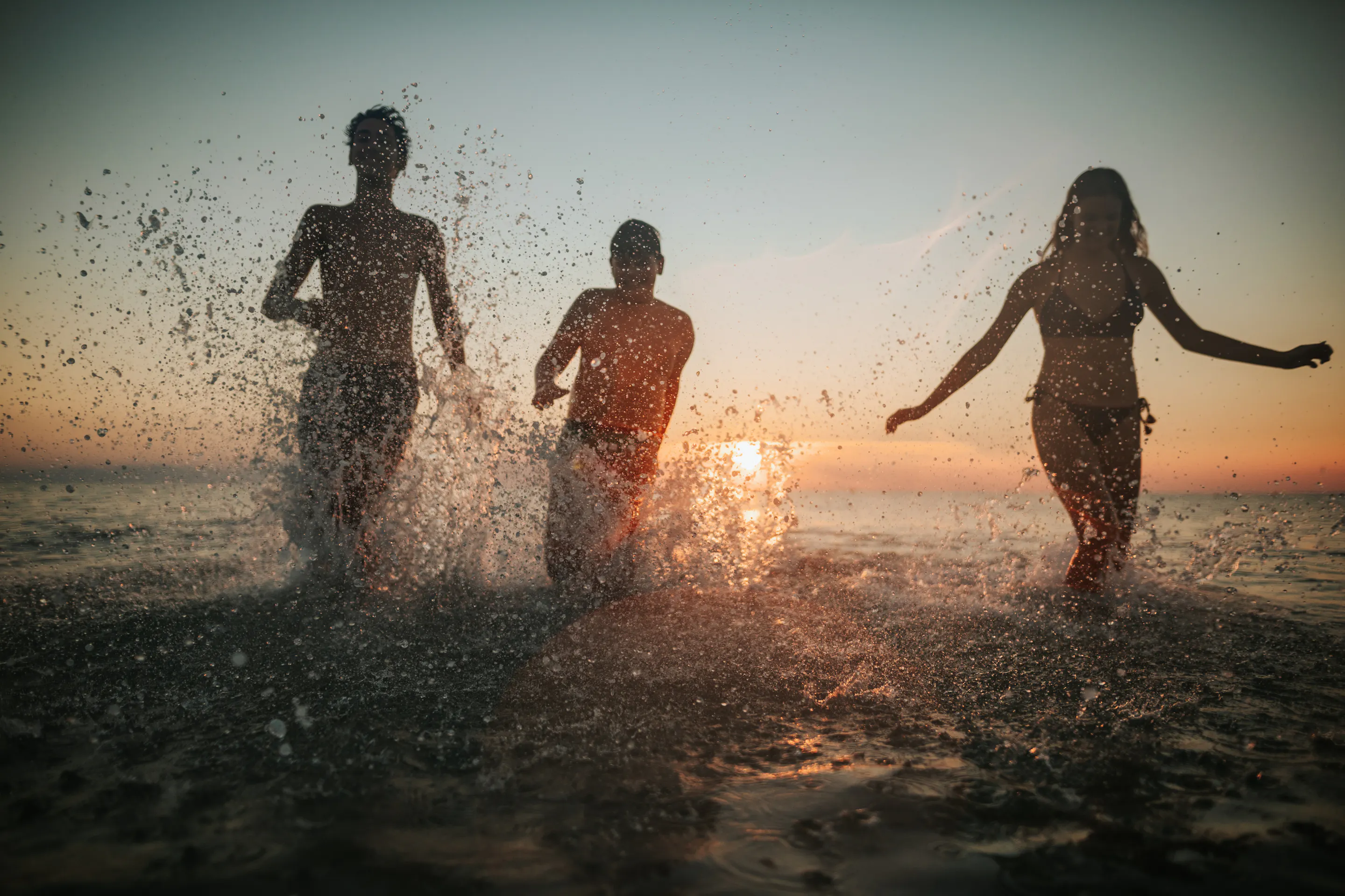 Three people running through shallow ocean water at sunset, splashing and backlit by golden light.