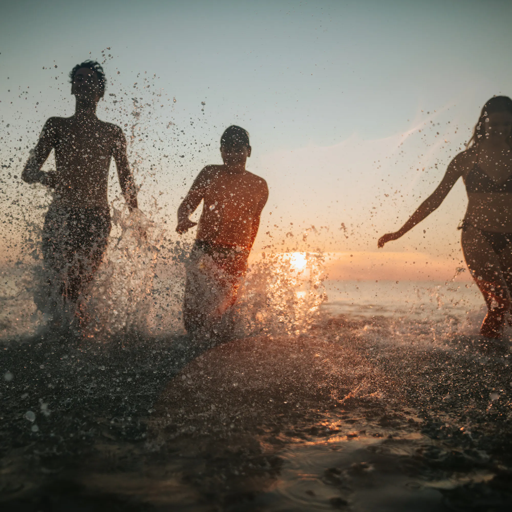 Three people running through shallow ocean water at sunset, splashing and backlit by golden light.