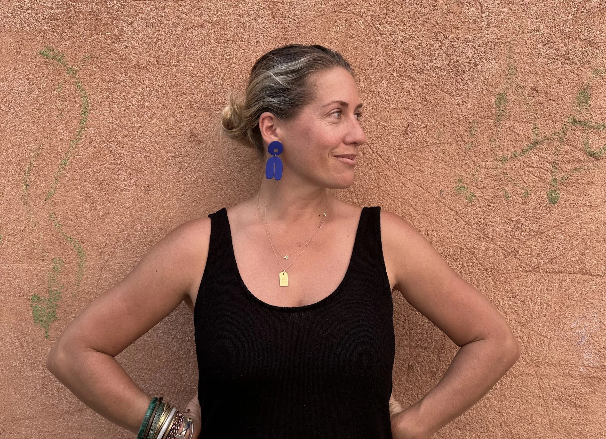 Woman in a black sleeveless top standing against a textured terracotta wall, looking to the side with hands on hips.