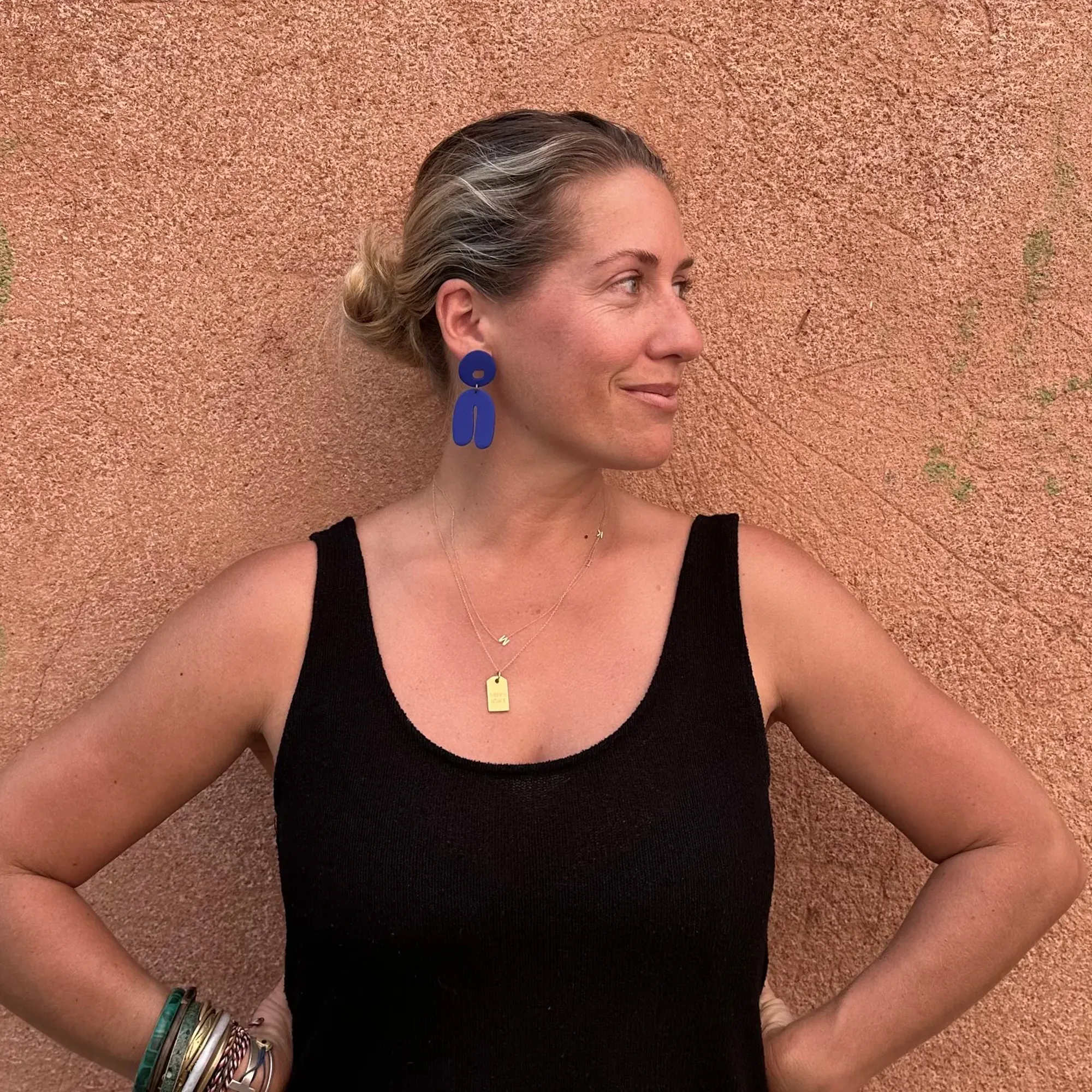 Woman in a black sleeveless top standing against a textured terracotta wall, looking to the side with hands on hips.