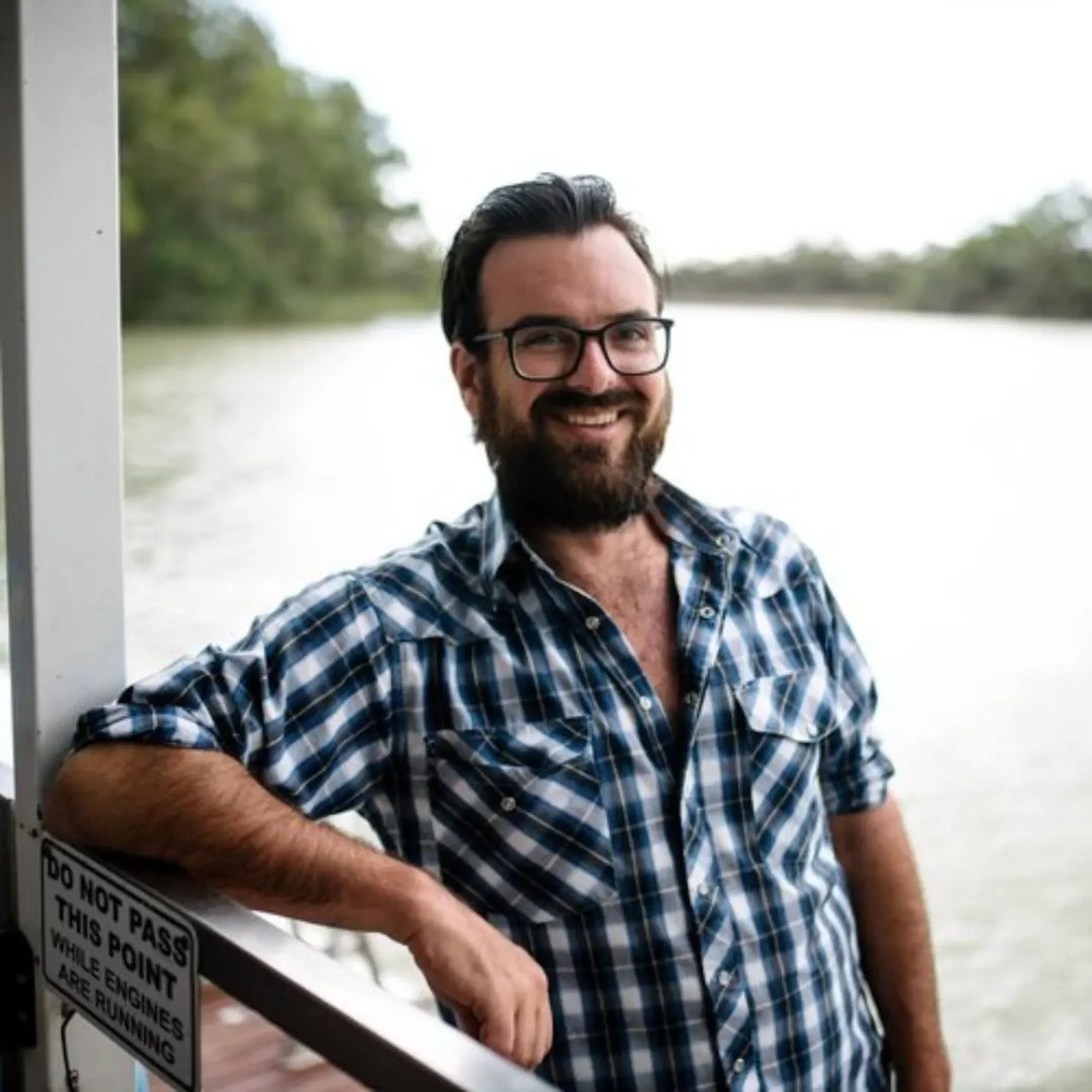 Man with glasses and a beard smiling while leaning on a railing by a river, wearing a blue plaid shirt.