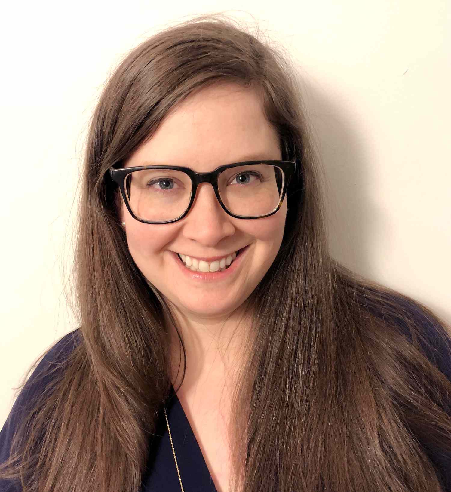 Woman with long brown hair and glasses smiling at the camera against a plain light background.