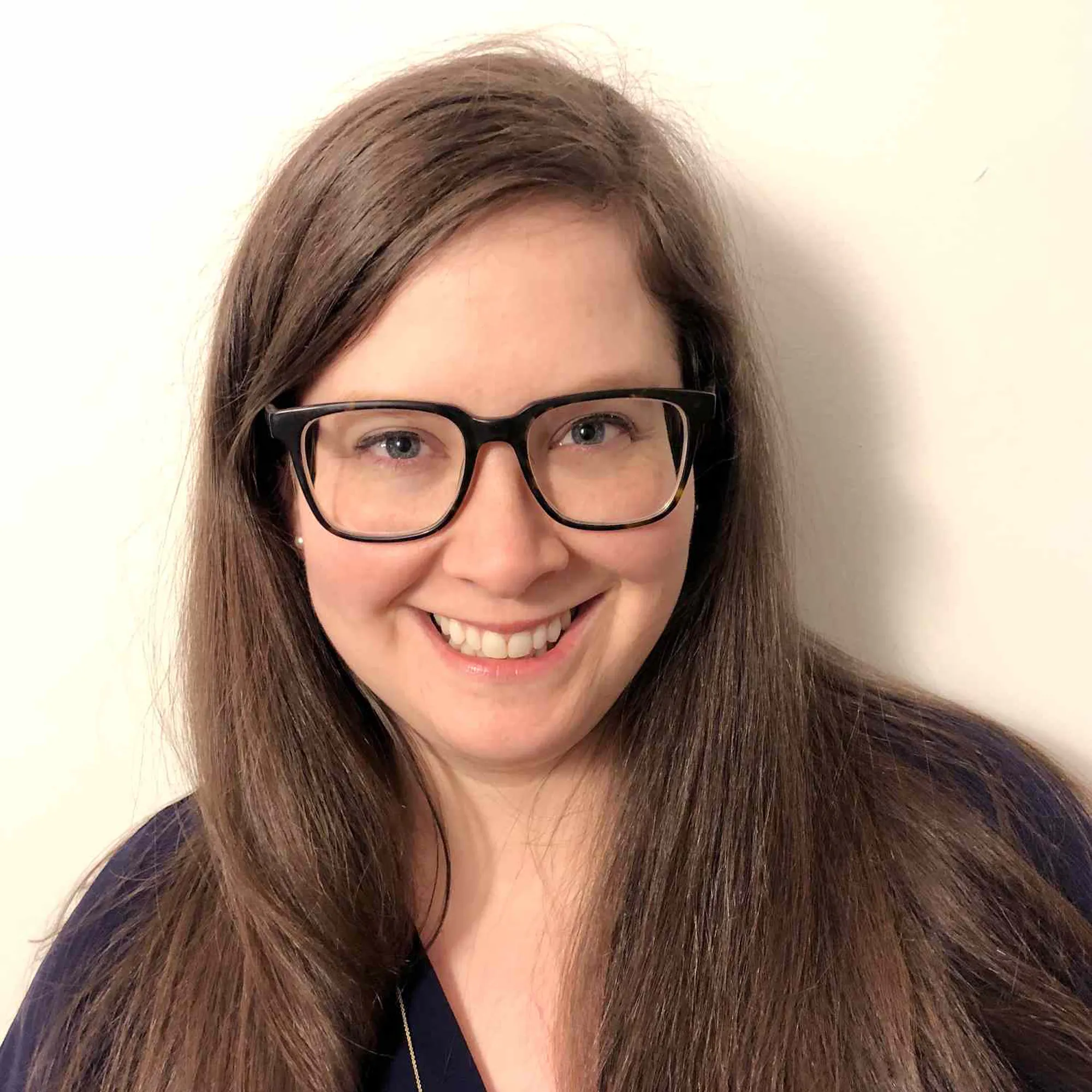 Woman with long brown hair and glasses smiling at the camera against a plain light background.