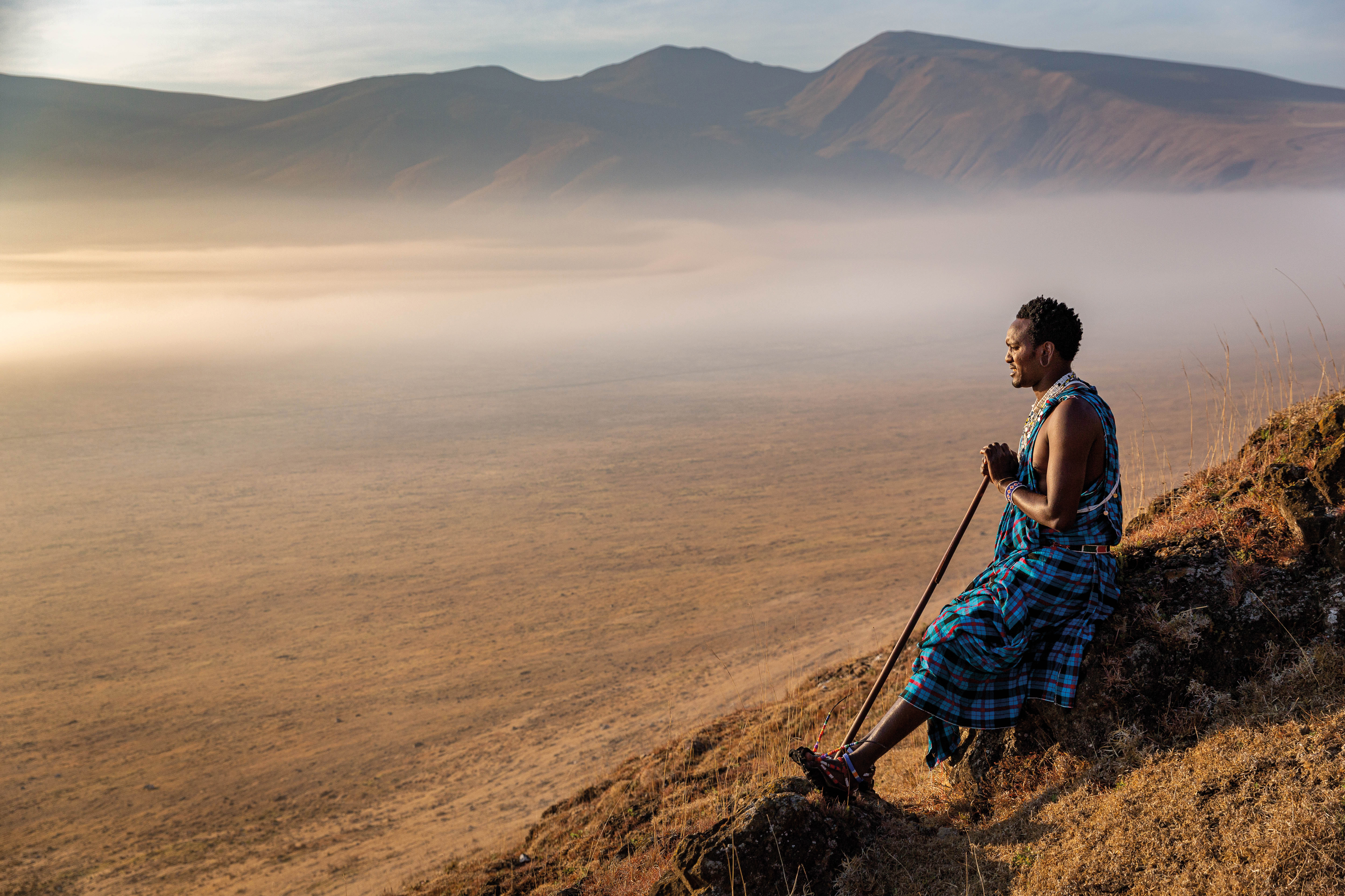 Maasai man in traditional clothing standing on a hillside, looking out over a vast, hazy savannah landscape.