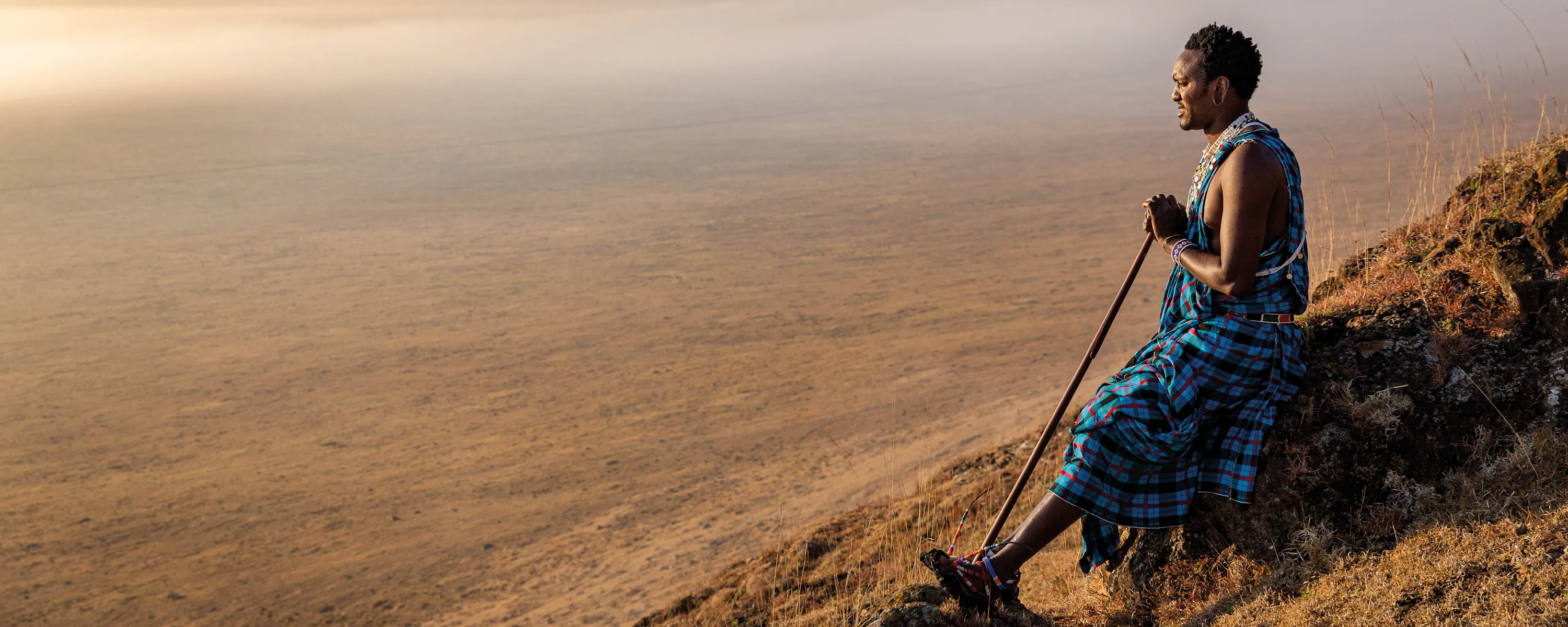 Maasai man in traditional clothing standing on a hillside, looking out over a vast, hazy savannah landscape.