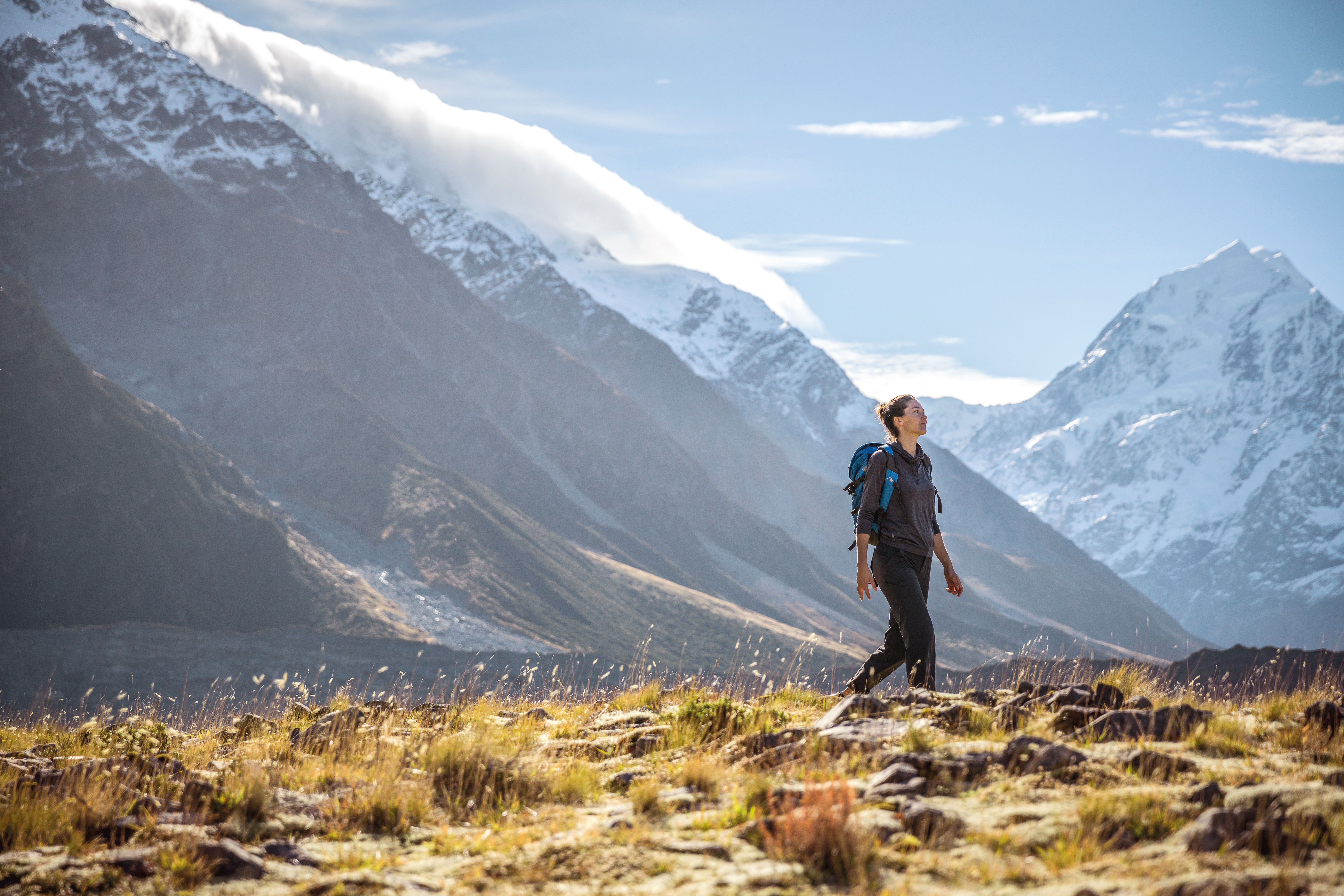 Woman with a backpack walking across a grassy ridge with dramatic snow-capped mountains and blue sky in the background.