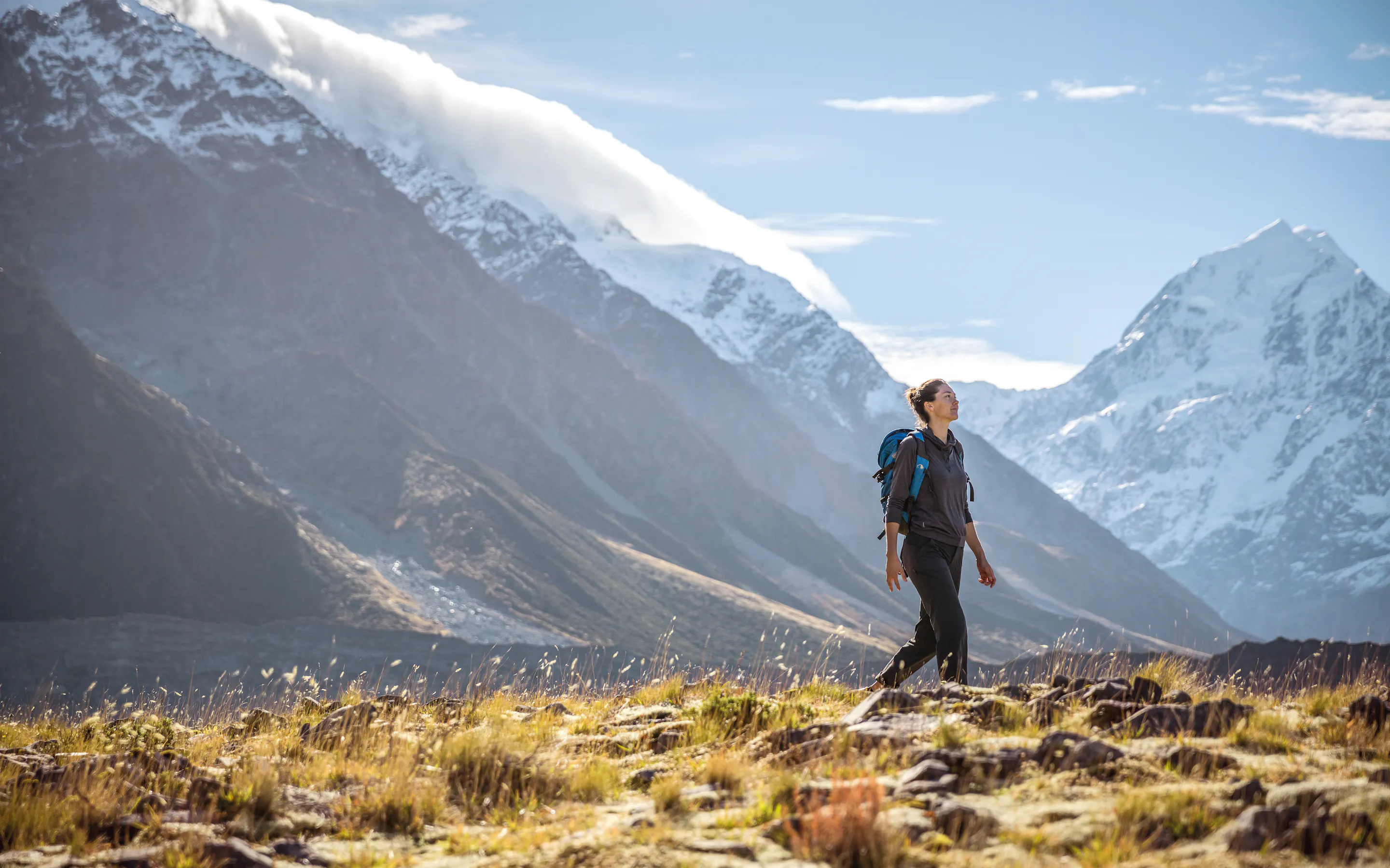 Woman with a backpack walking across a grassy ridge with dramatic snow-capped mountains and blue sky in the background.