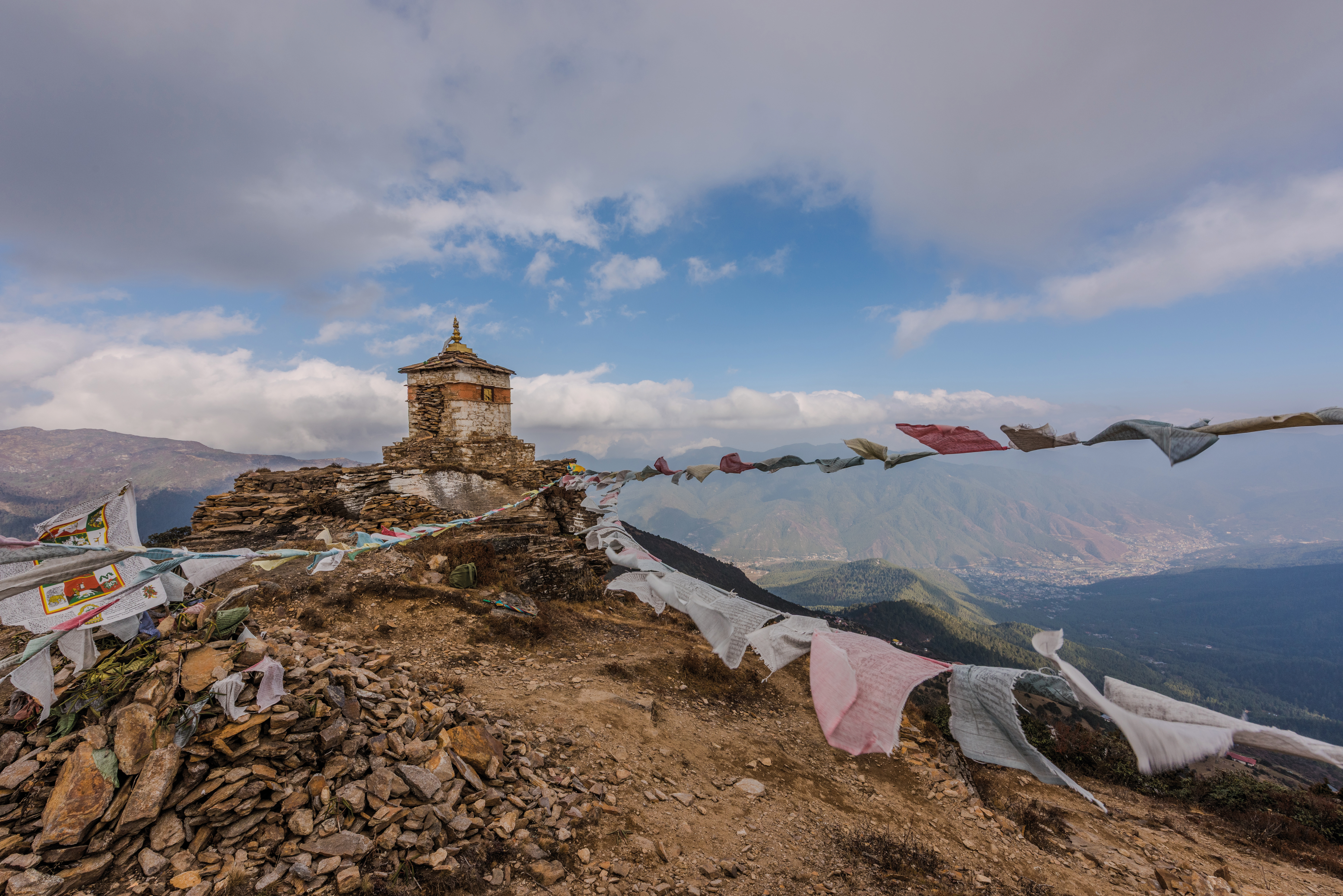 Small stone shrine on a mountain ridge with colorful prayer flags blowing in the wind and distant hills beyond.