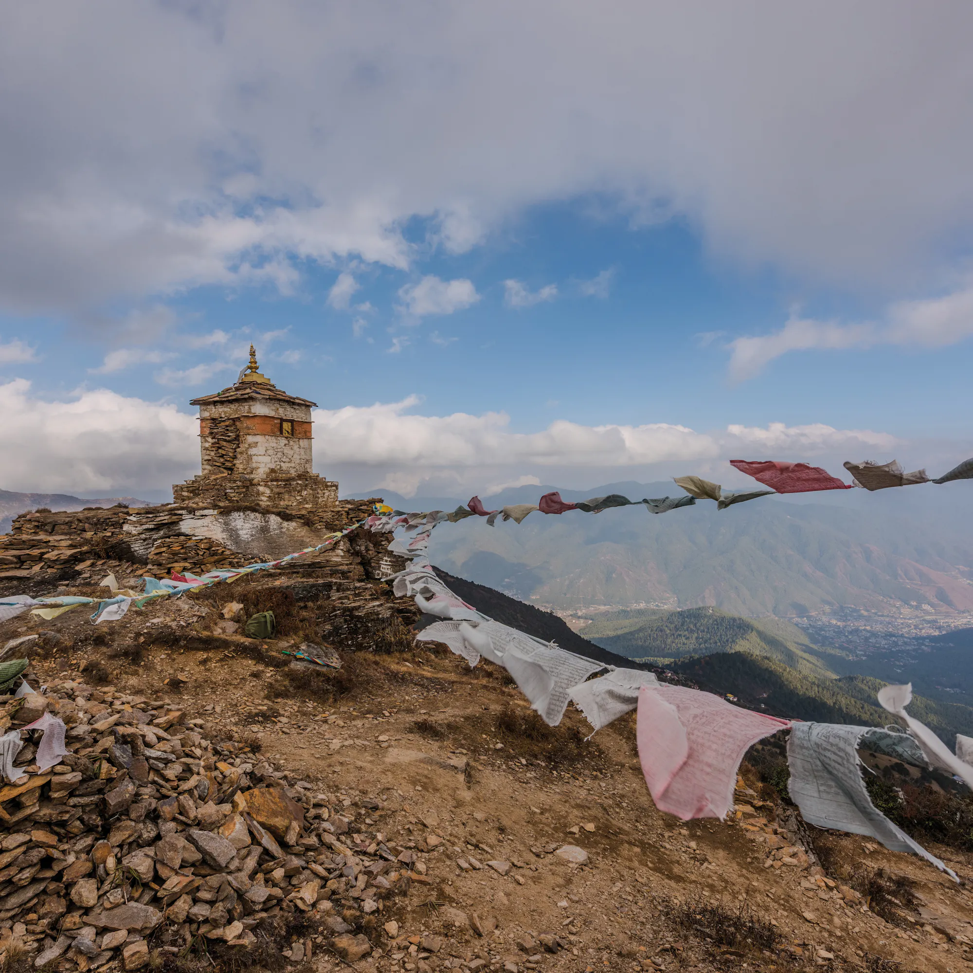 Small stone shrine on a mountain ridge with colorful prayer flags blowing in the wind and distant hills beyond.