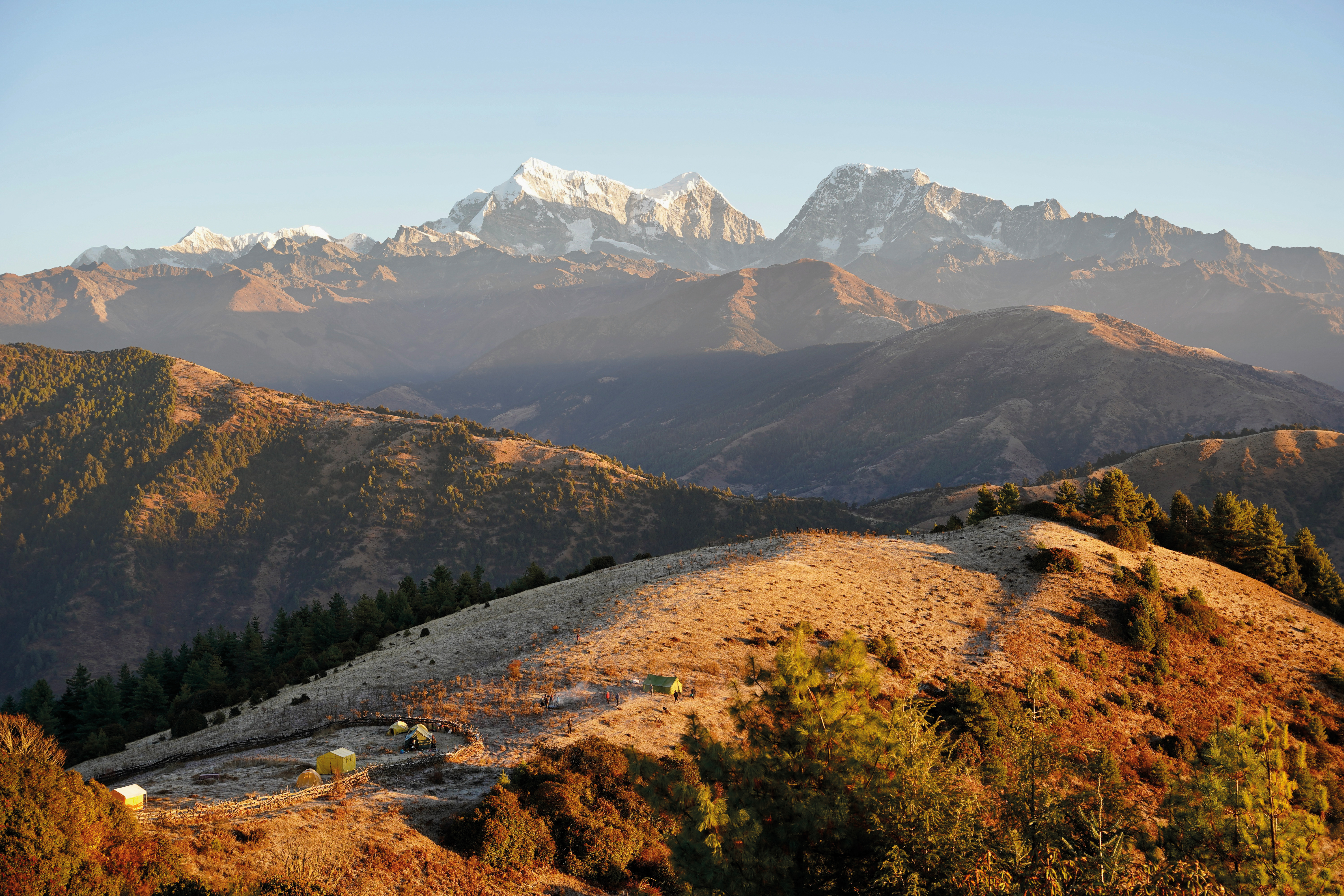 Rolling hills with a small campsite and tents, backed by a range of snow-capped mountains in warm early light.