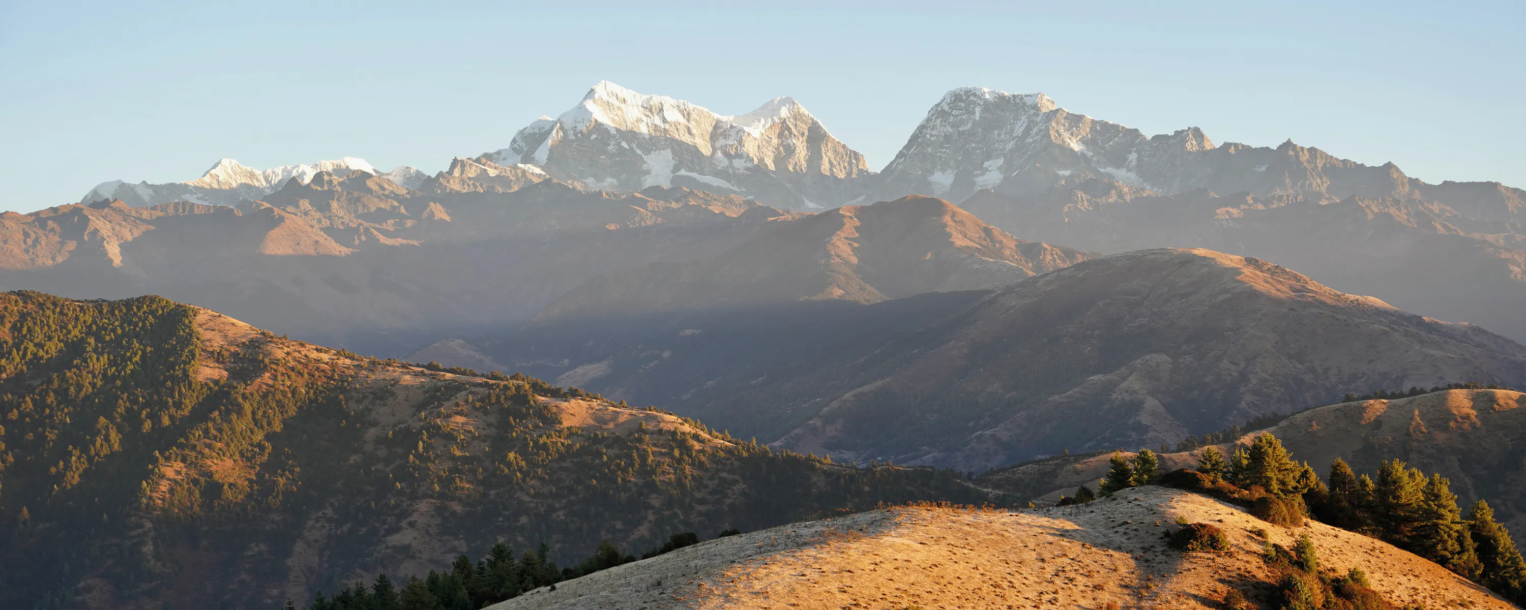 Rolling hills with a small campsite and tents, backed by a range of snow-capped mountains in warm early light.