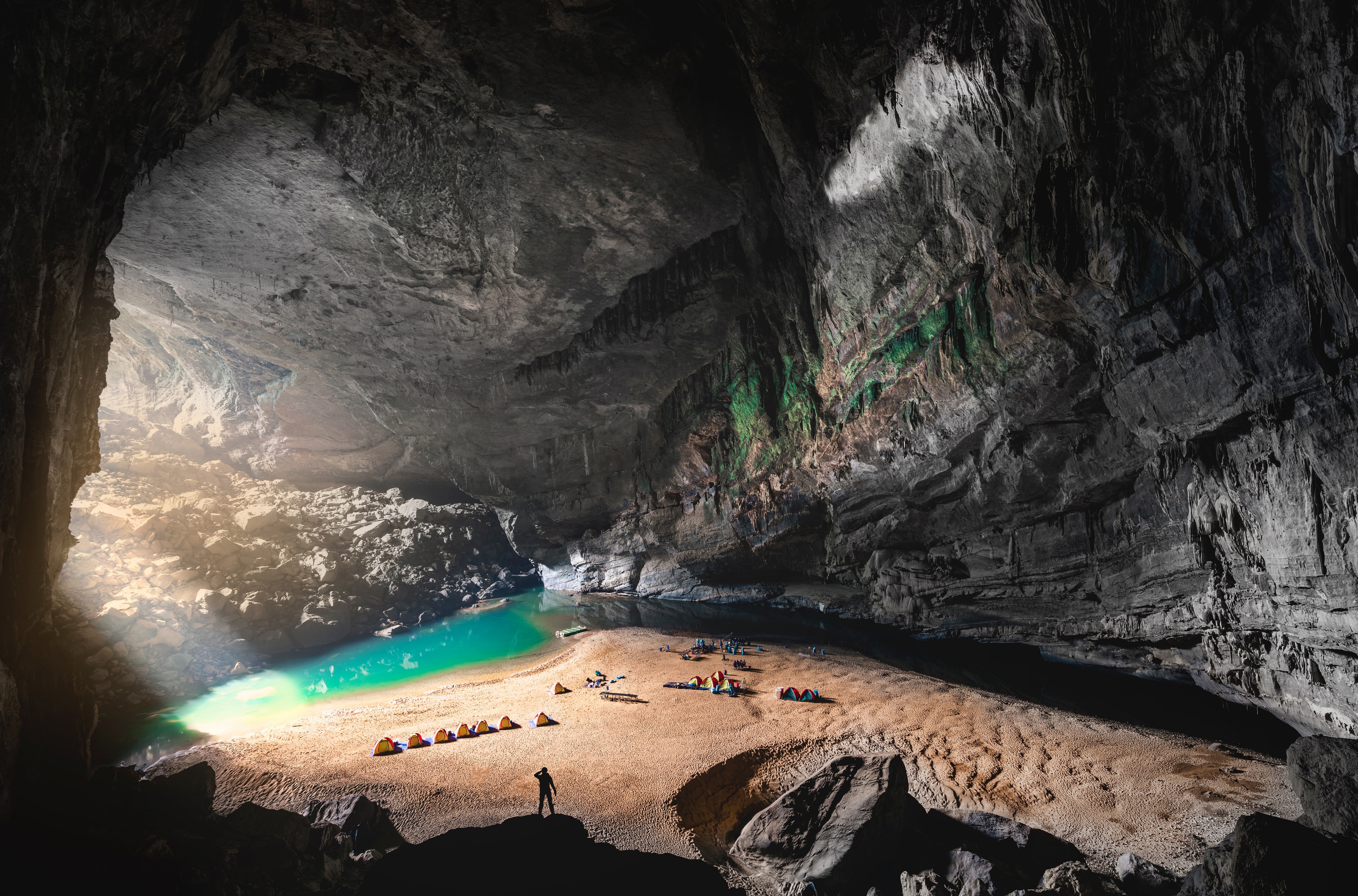 Tiny tents line a sandy bank beside a blue lagoon inside Son Doong Cave, under a vast rock ceiling lit from above.