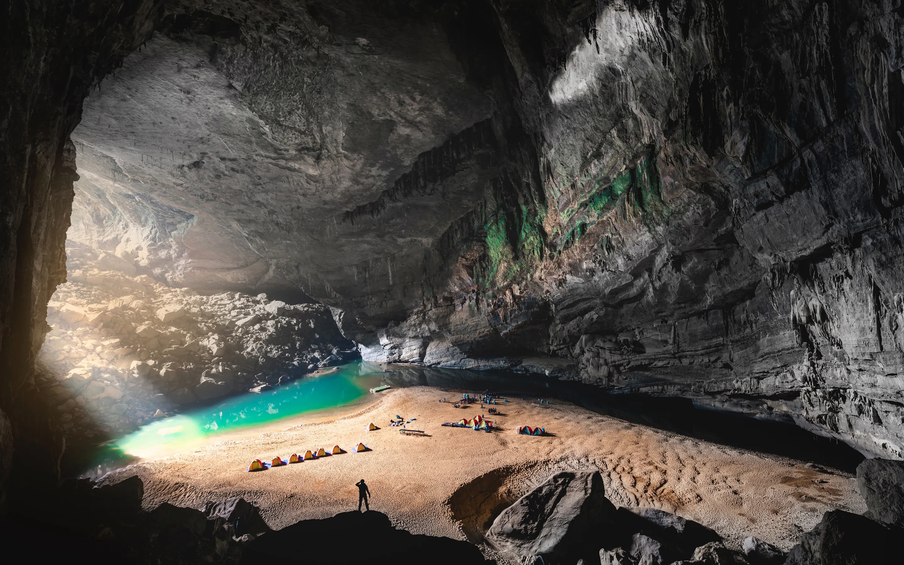 Tiny tents line a sandy bank beside a blue lagoon inside Son Doong Cave, under a vast rock ceiling lit from above.
