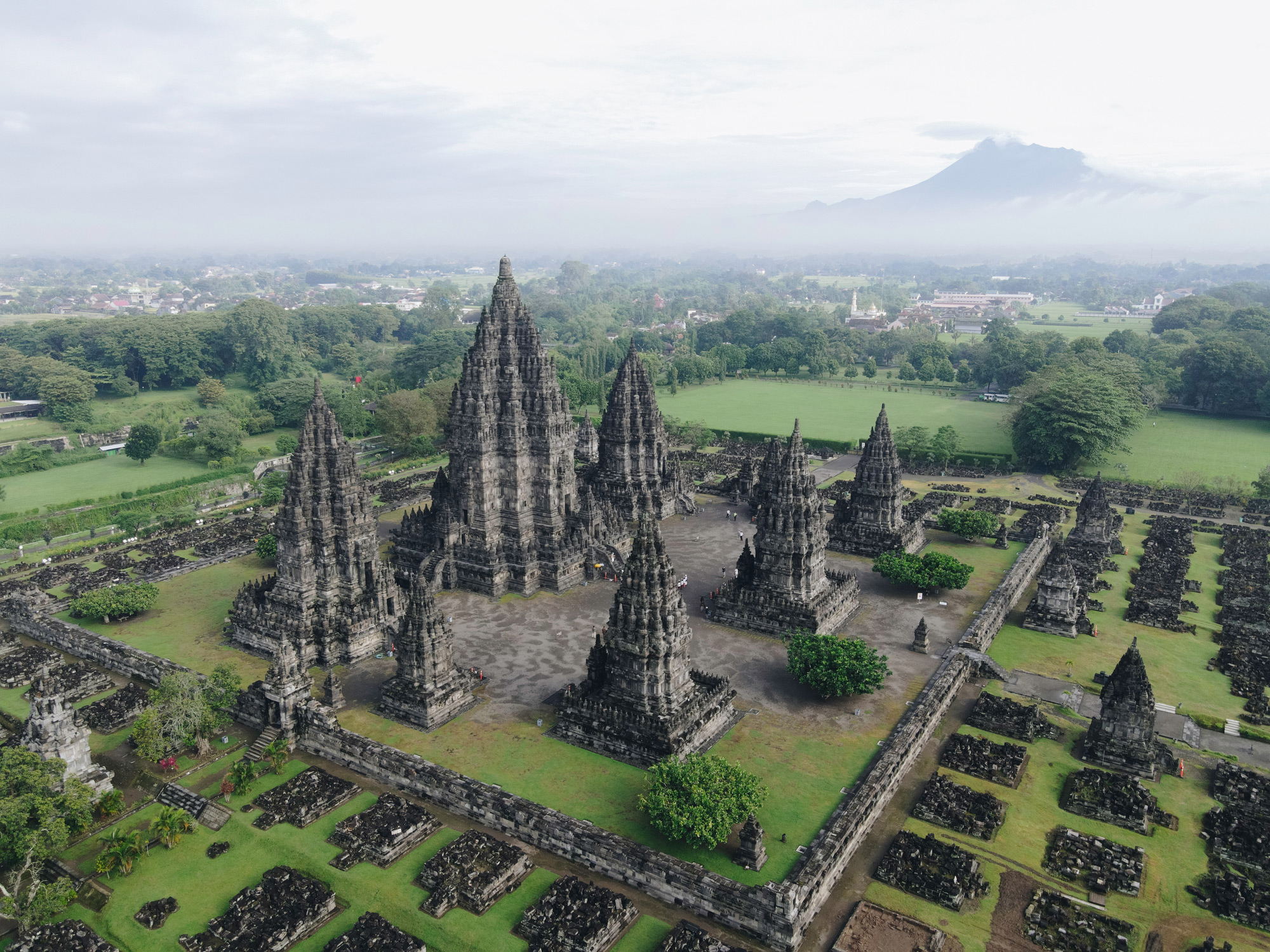 An aerial view shows Prambanan's stone temple spires rising from green lawns beneath a pale sky in central Java.