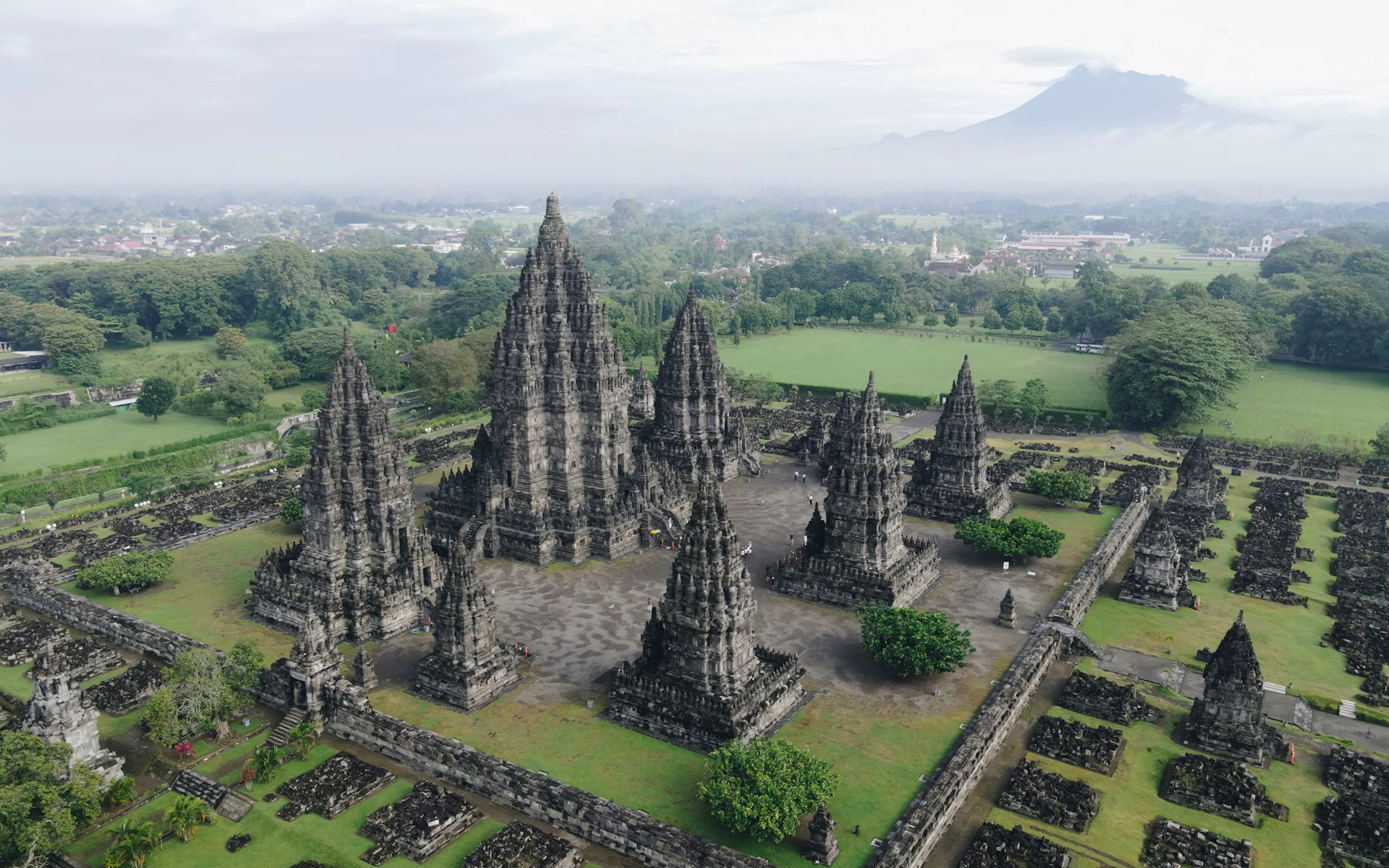 An aerial view shows Prambanan's stone temple spires rising from green lawns beneath a pale sky in central Java.