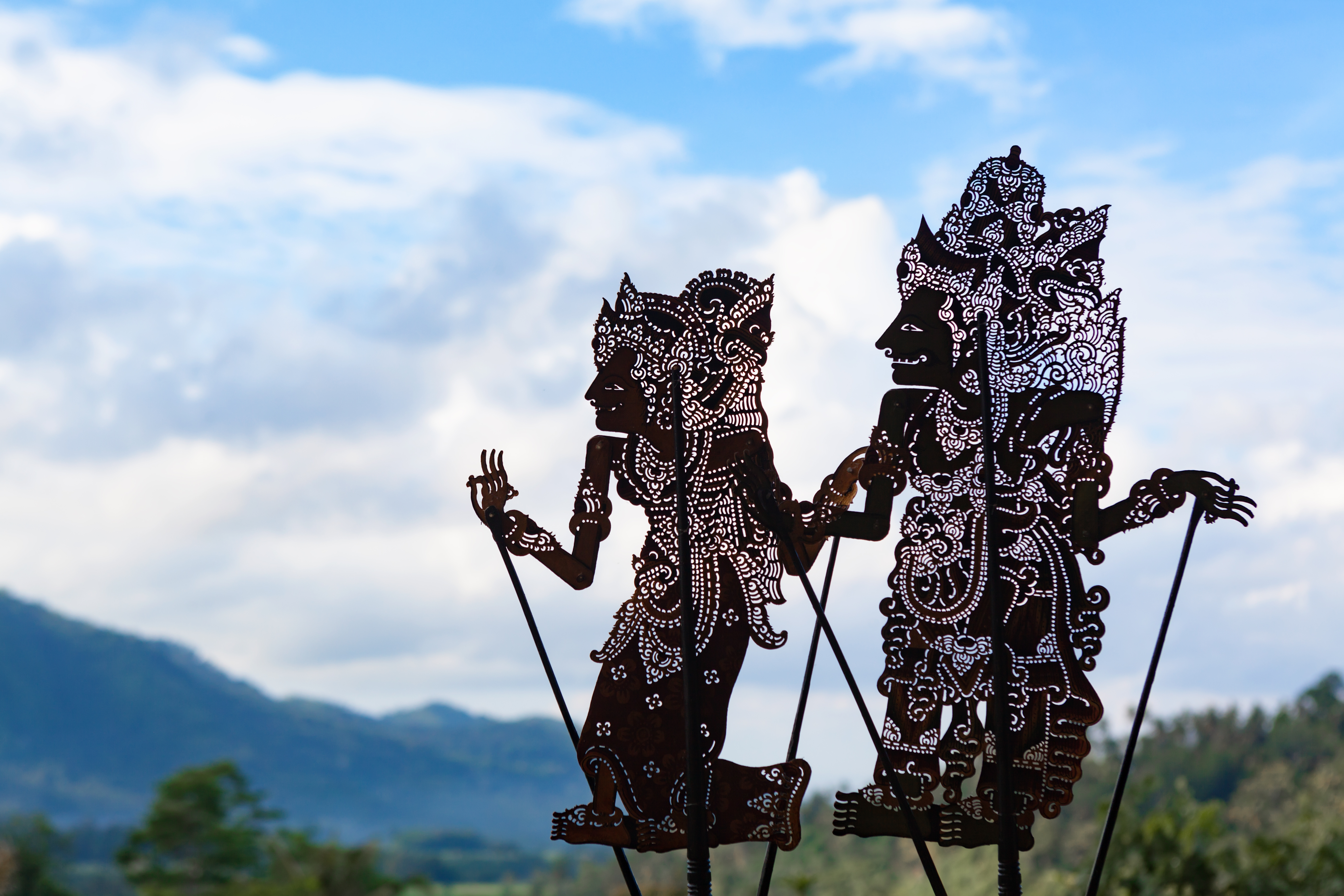 Two shadow puppets stand against a soft blue sky in Bali, their cutout patterns and carved profiles sharply defined.