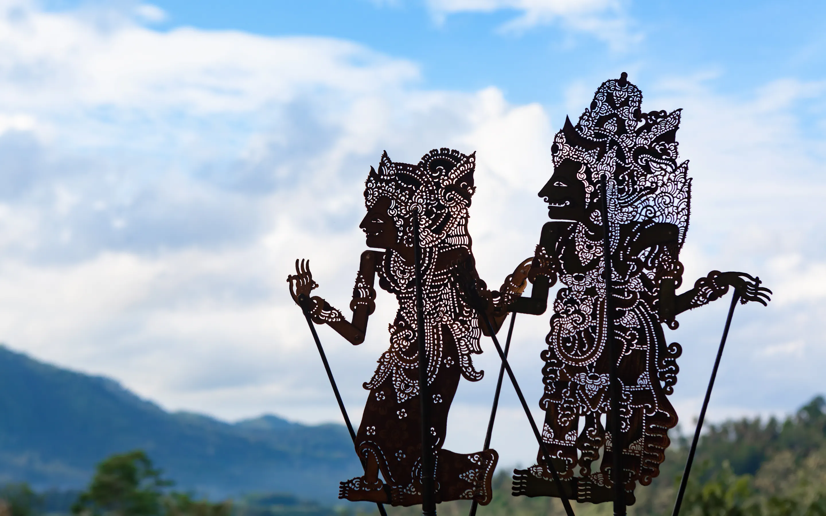 Two shadow puppets stand against a soft blue sky in Bali, their cutout patterns and carved profiles sharply defined.