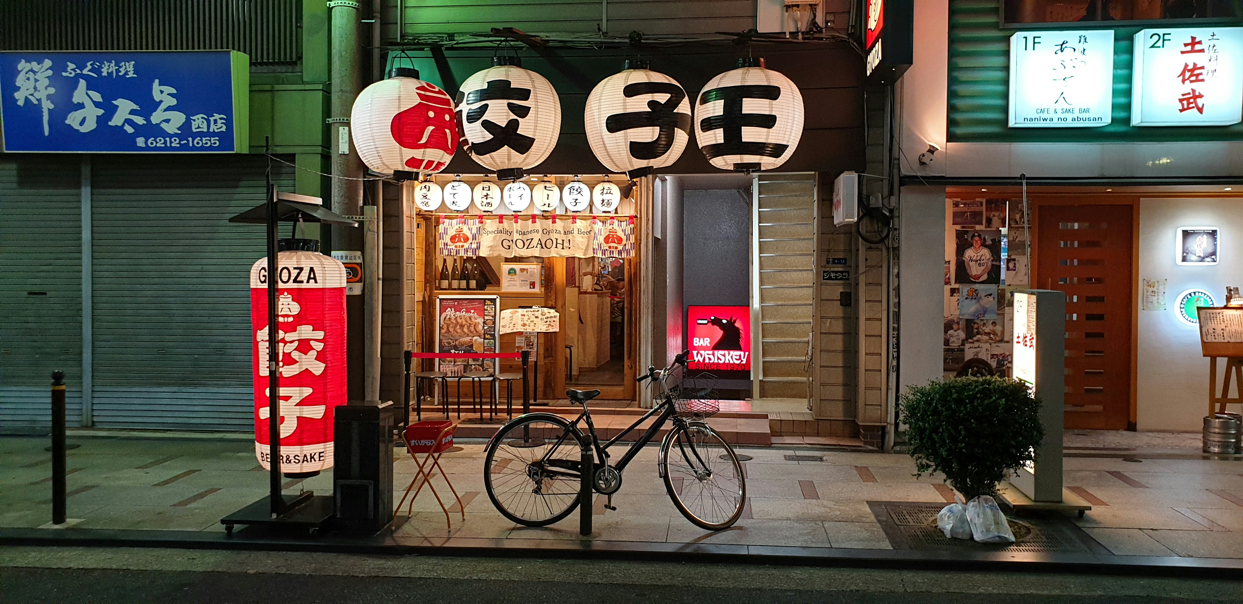 A bicycle stands outside a bright gyoza shop in Osaka at night, framed by lanterns, signs, and neighboring storefronts.