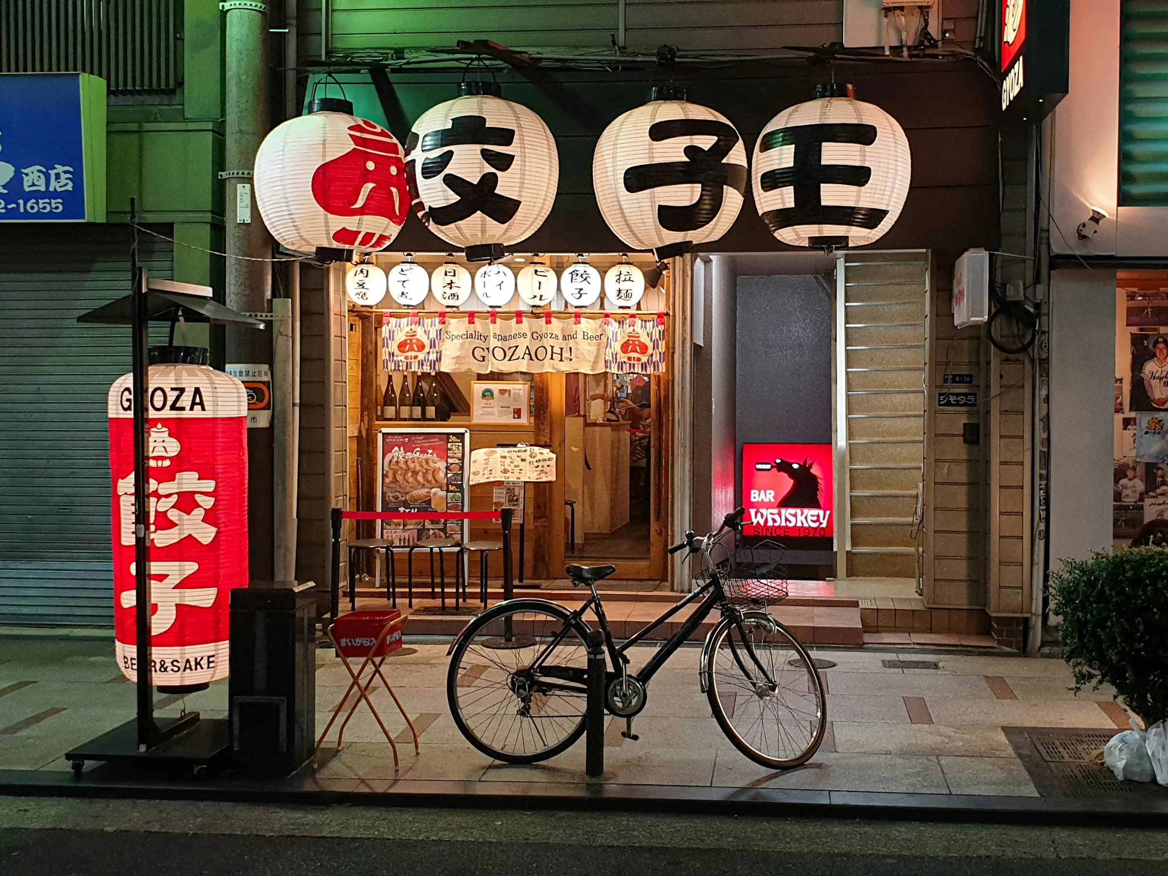 A bicycle stands outside a bright gyoza shop in Osaka at night, framed by lanterns, signs, and neighboring storefronts.