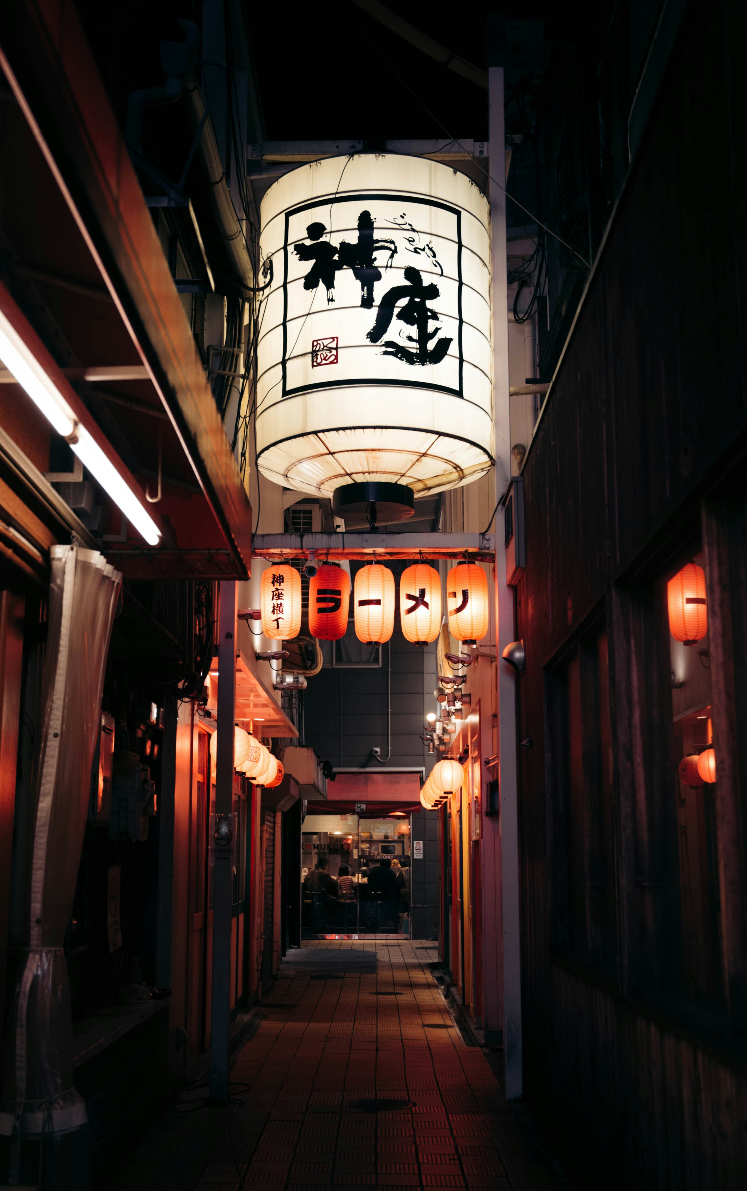 A glowing paper lantern hangs above a narrow Osaka alley at night, with red shop lights and dark doorways receding behind.