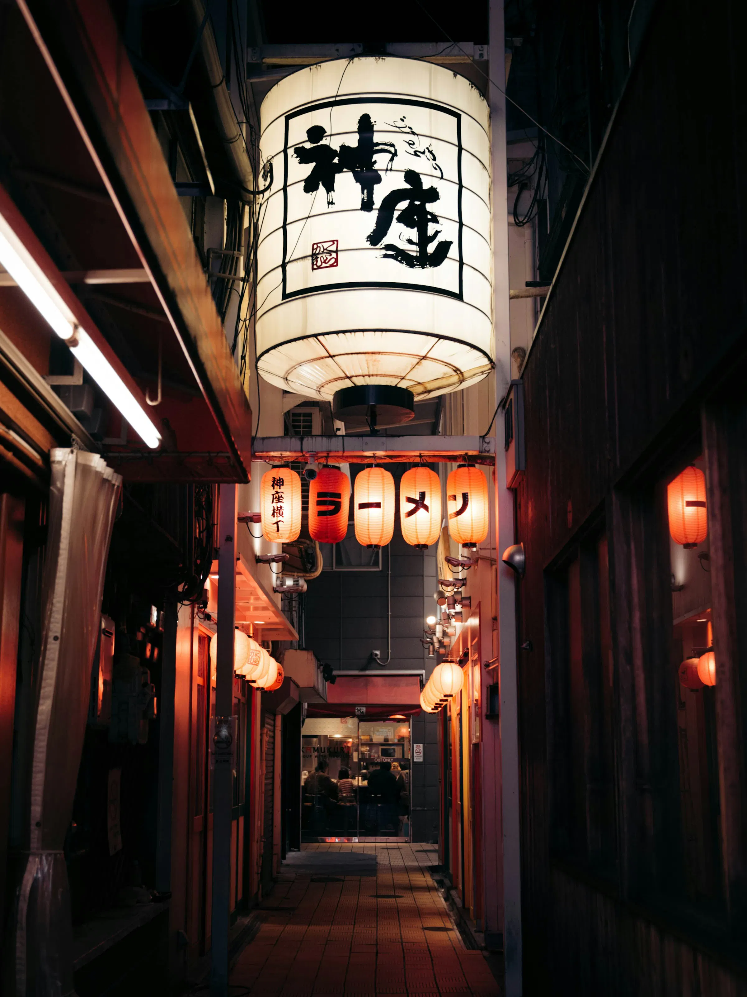 A glowing paper lantern hangs above a narrow Osaka alley at night, with red shop lights and dark doorways receding behind.