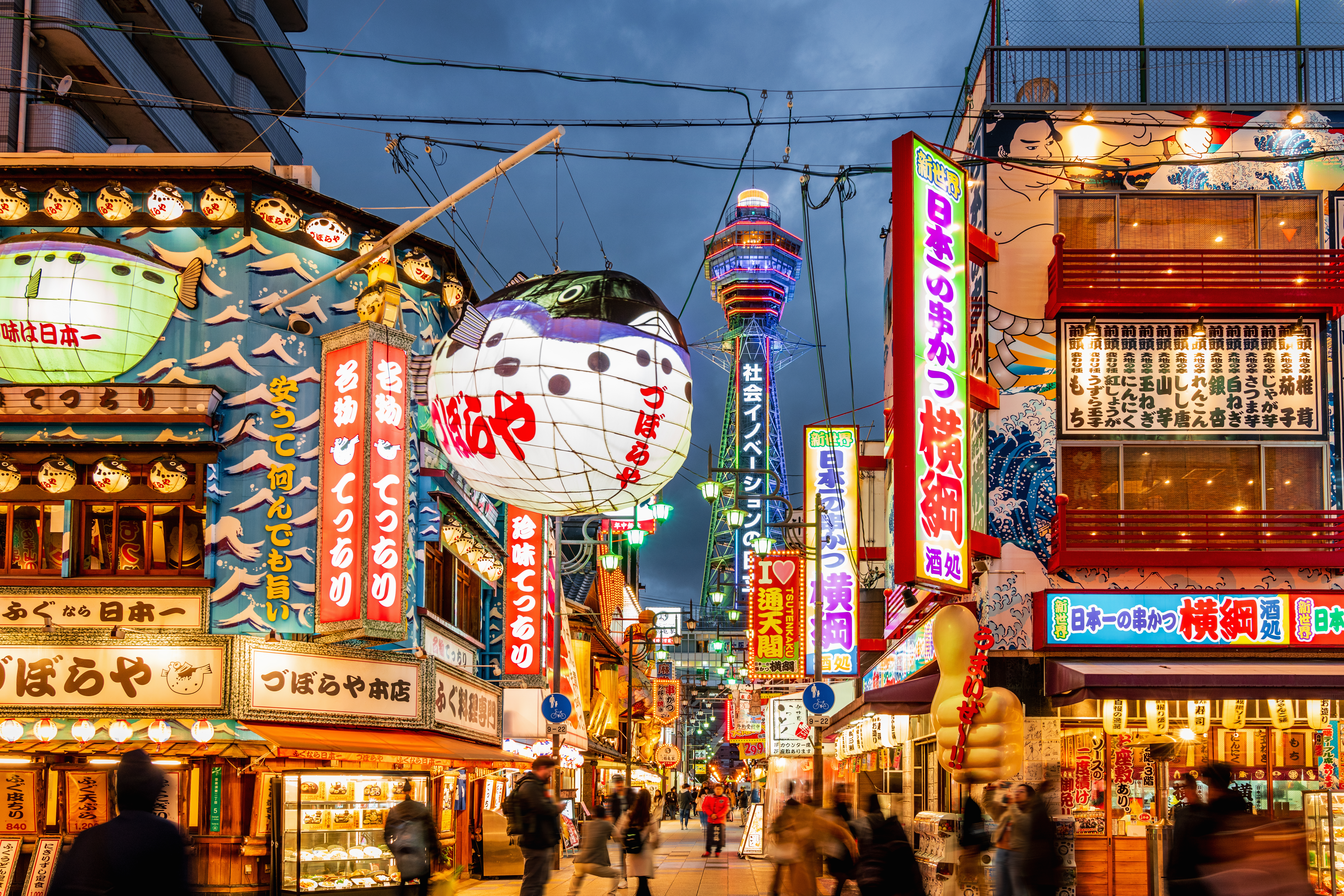 Bright neon signs and busy streets fill Osaka's Shinsekai district at dusk, with Tsutenkaku Tower rising behind.