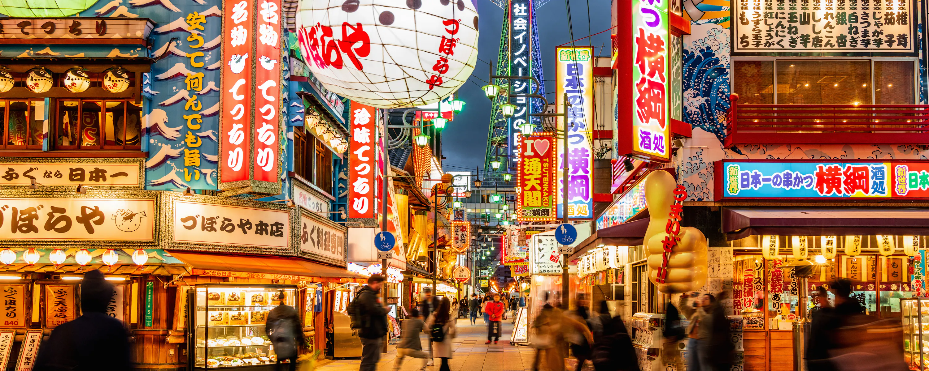 Bright neon signs and busy streets fill Osaka's Shinsekai district at dusk, with Tsutenkaku Tower rising behind.