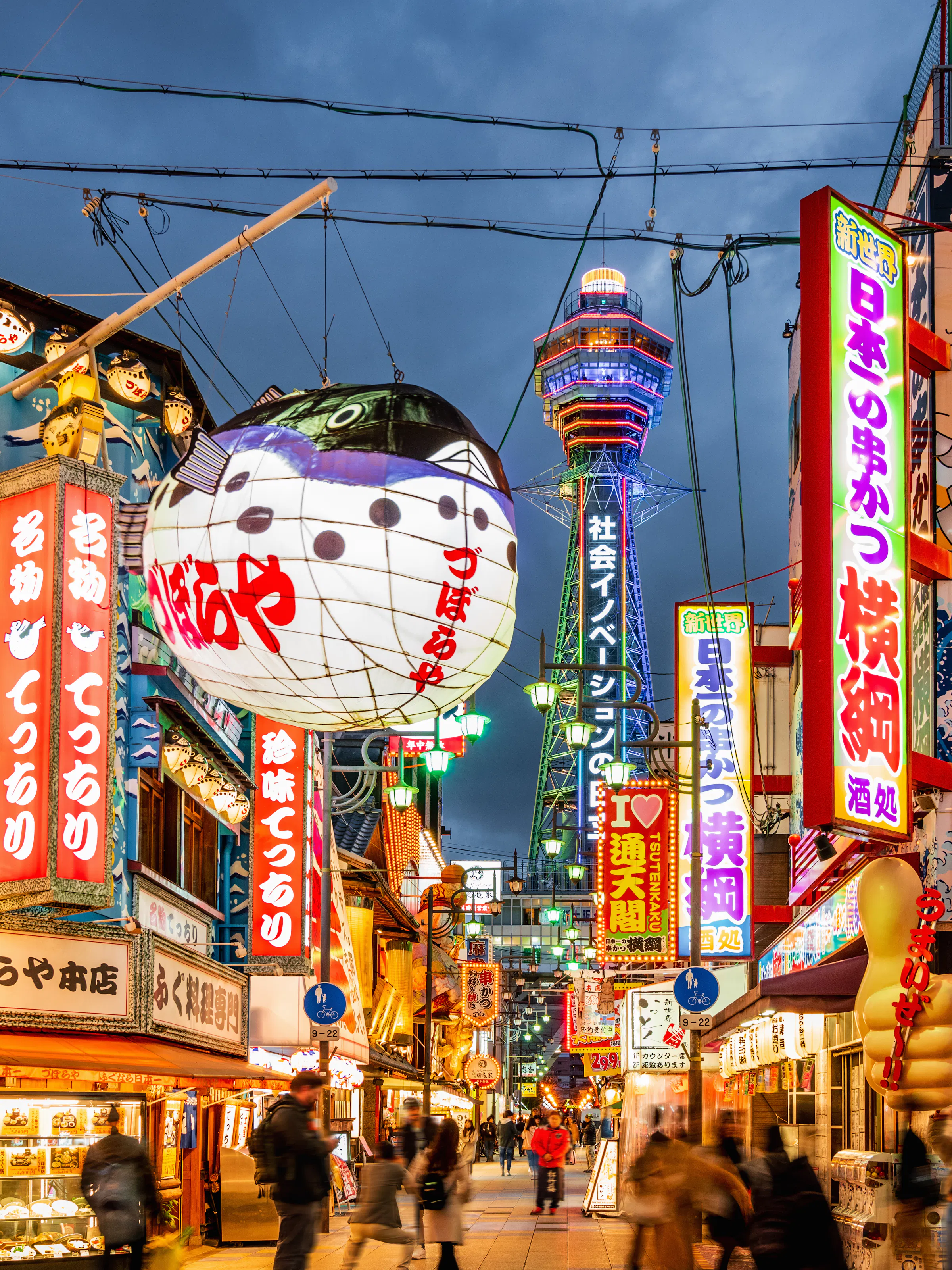 Bright neon signs and busy streets fill Osaka's Shinsekai district at dusk, with Tsutenkaku Tower rising behind.