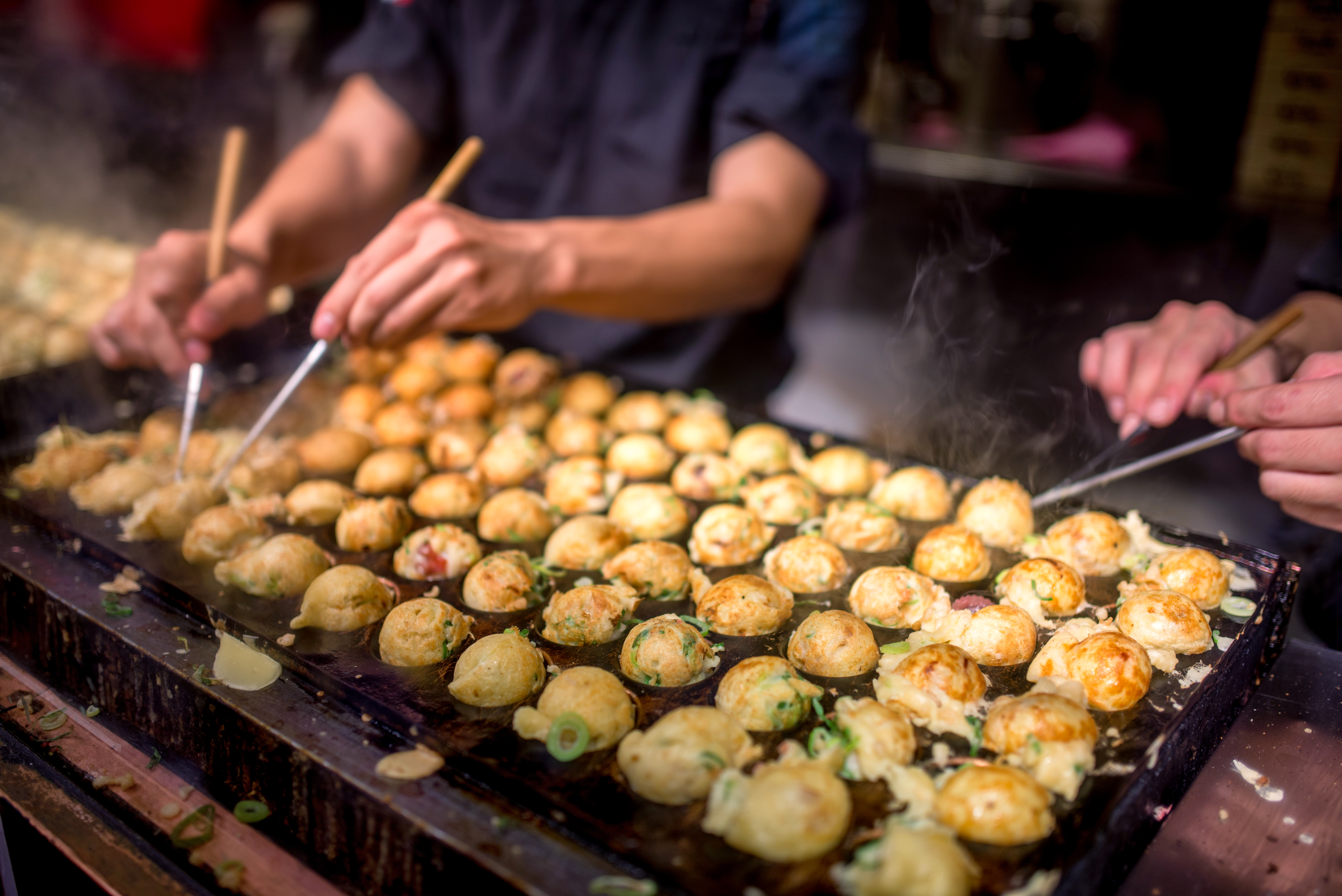 Rows of takoyaki cook on a hot grill as several hands turn the batter balls with picks at a busy Osaka food stall.