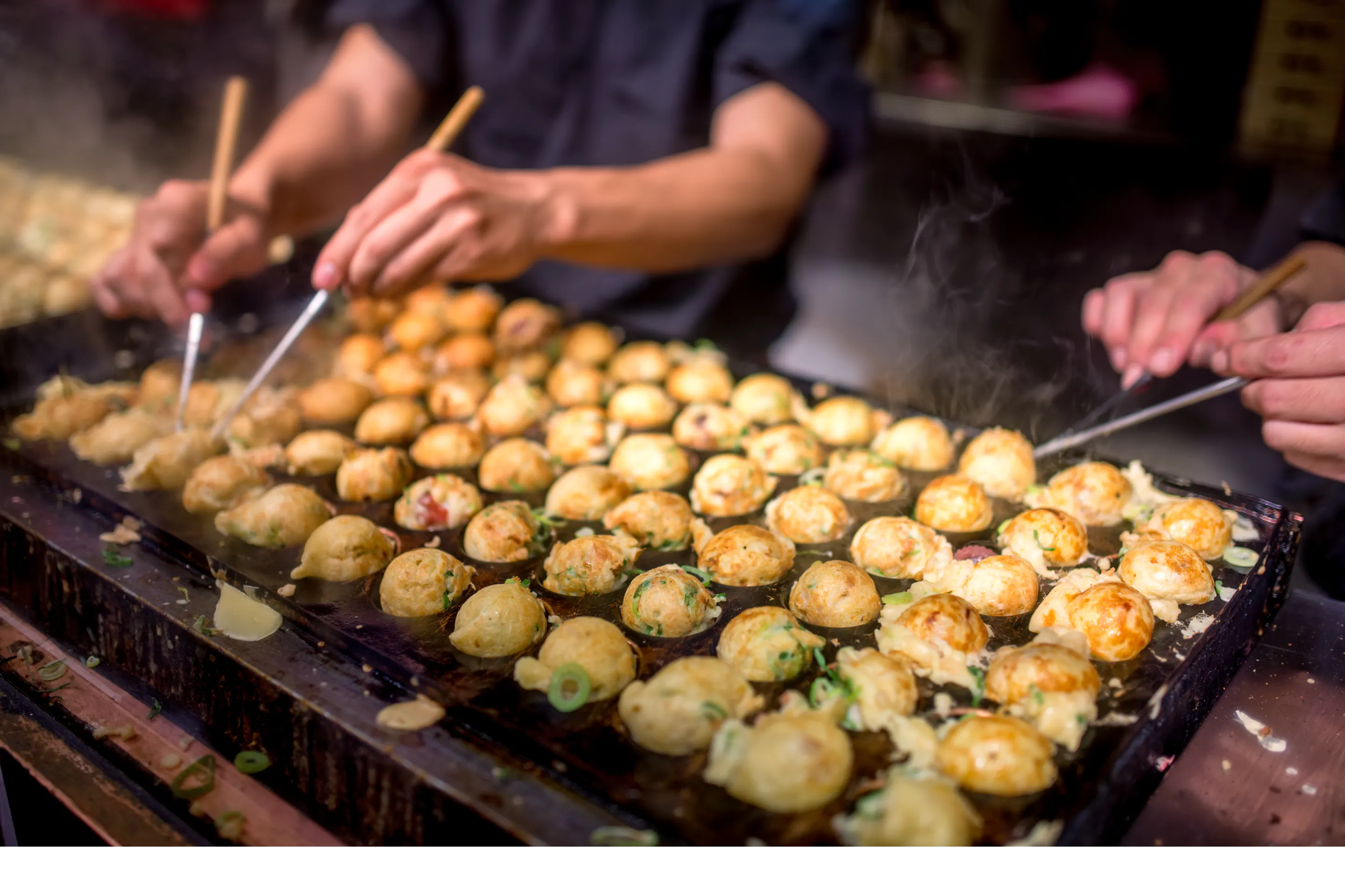 Rows of takoyaki cook on a hot grill as several hands turn the batter balls with picks at a busy Osaka food stall.