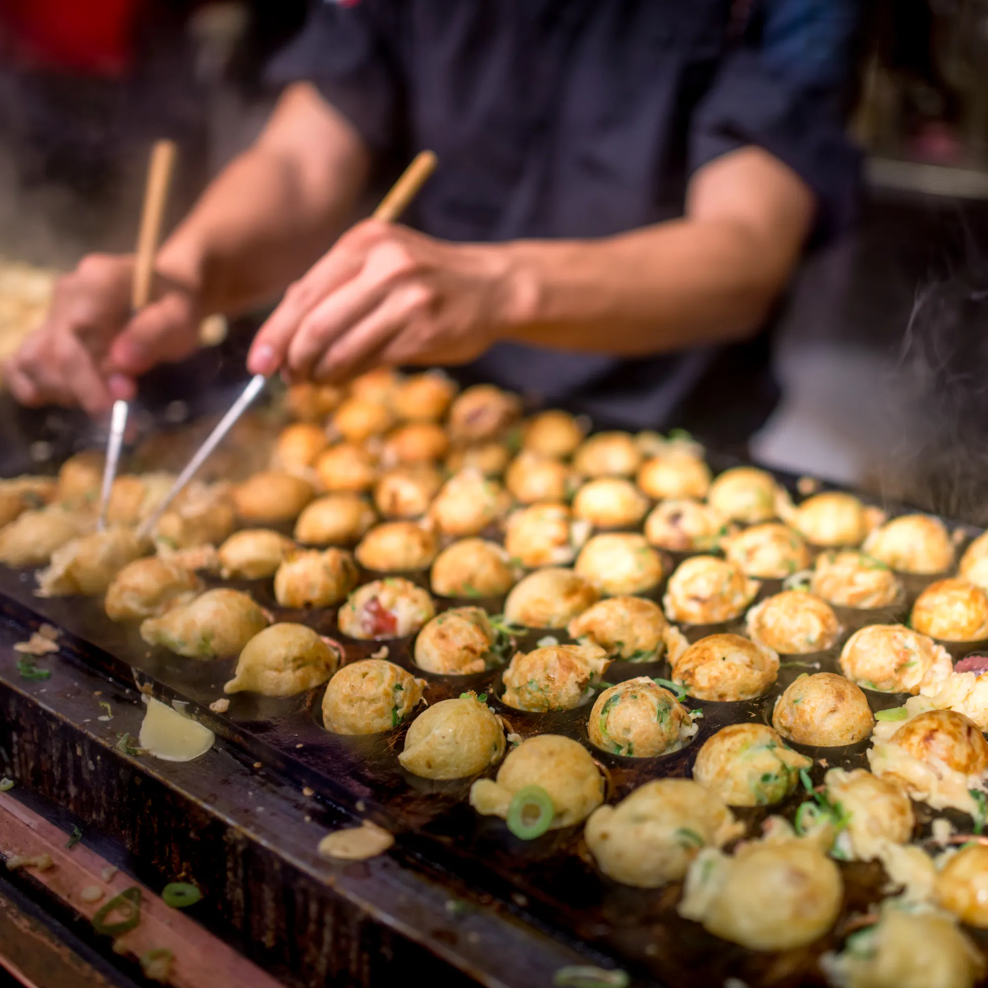 Rows of takoyaki cook on a hot grill as several hands turn the batter balls with picks at a busy Osaka food stall.