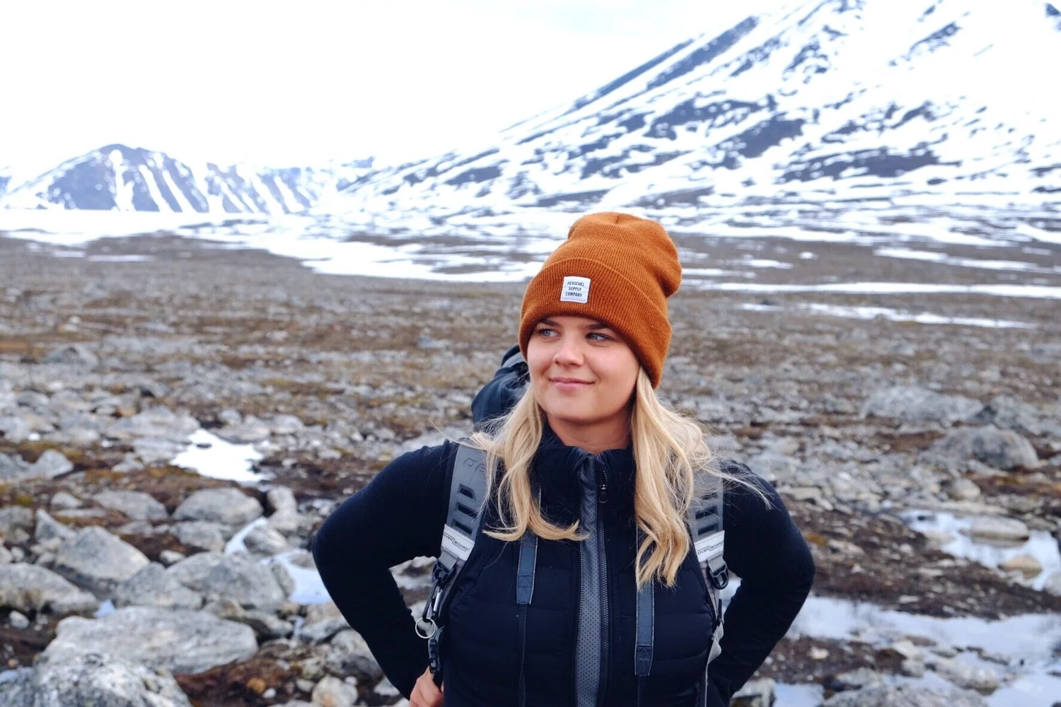 A woman with blonde hair wearing an orange beanie and a backpack stands in front of a snowy mountain.