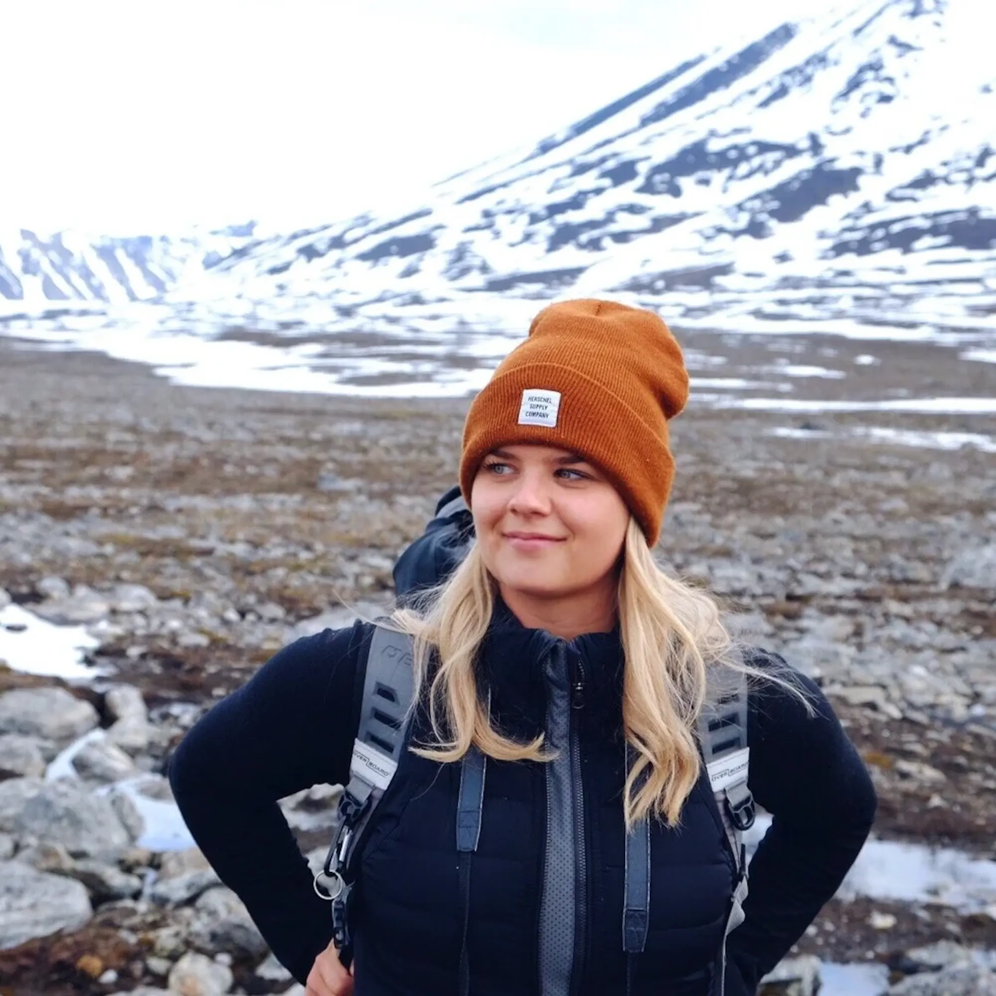 A woman with blonde hair wearing an orange beanie and a backpack stands in front of a snowy mountain.