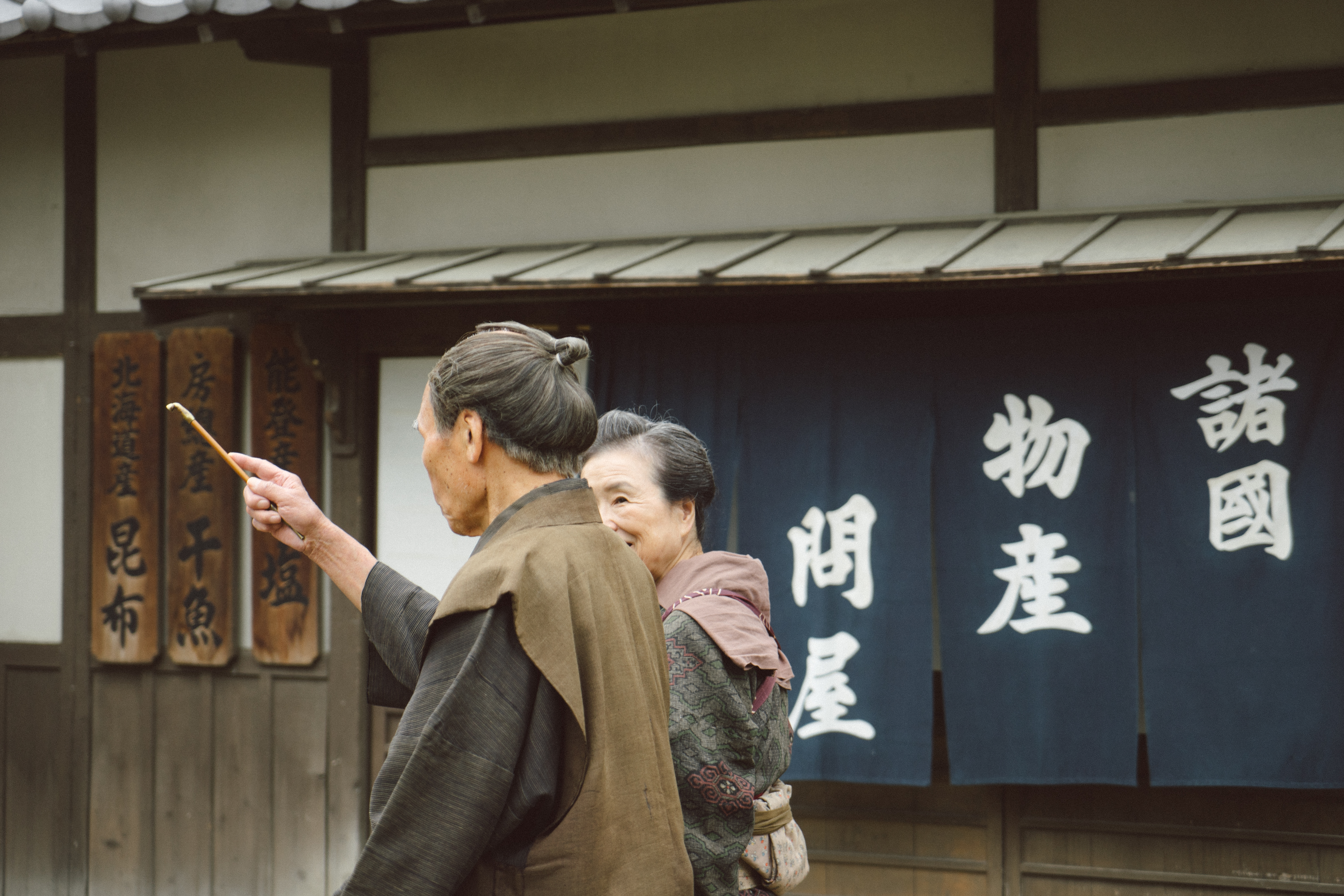 Two people in traditional dress stand beside a shopfront with bold Japanese signage in an Edo-style village setting.