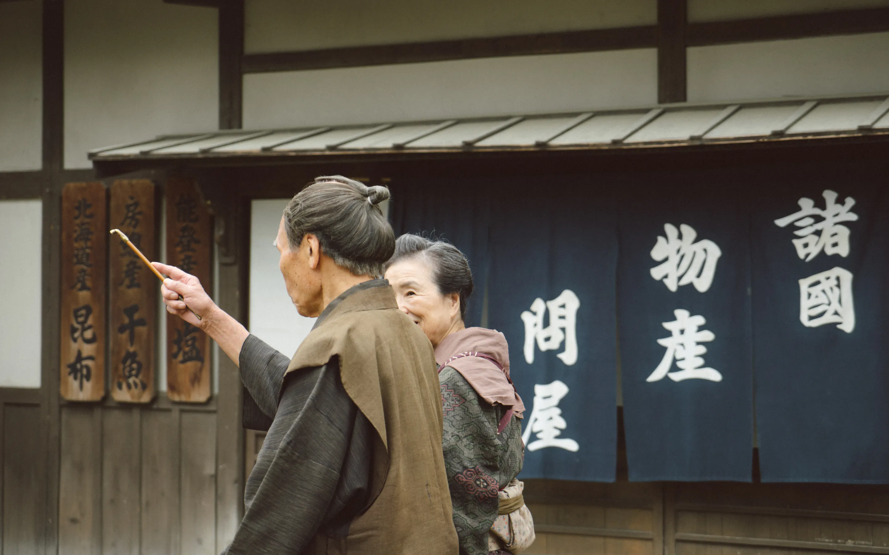 Two people in traditional dress stand beside a shopfront with bold Japanese signage in an Edo-style village setting.