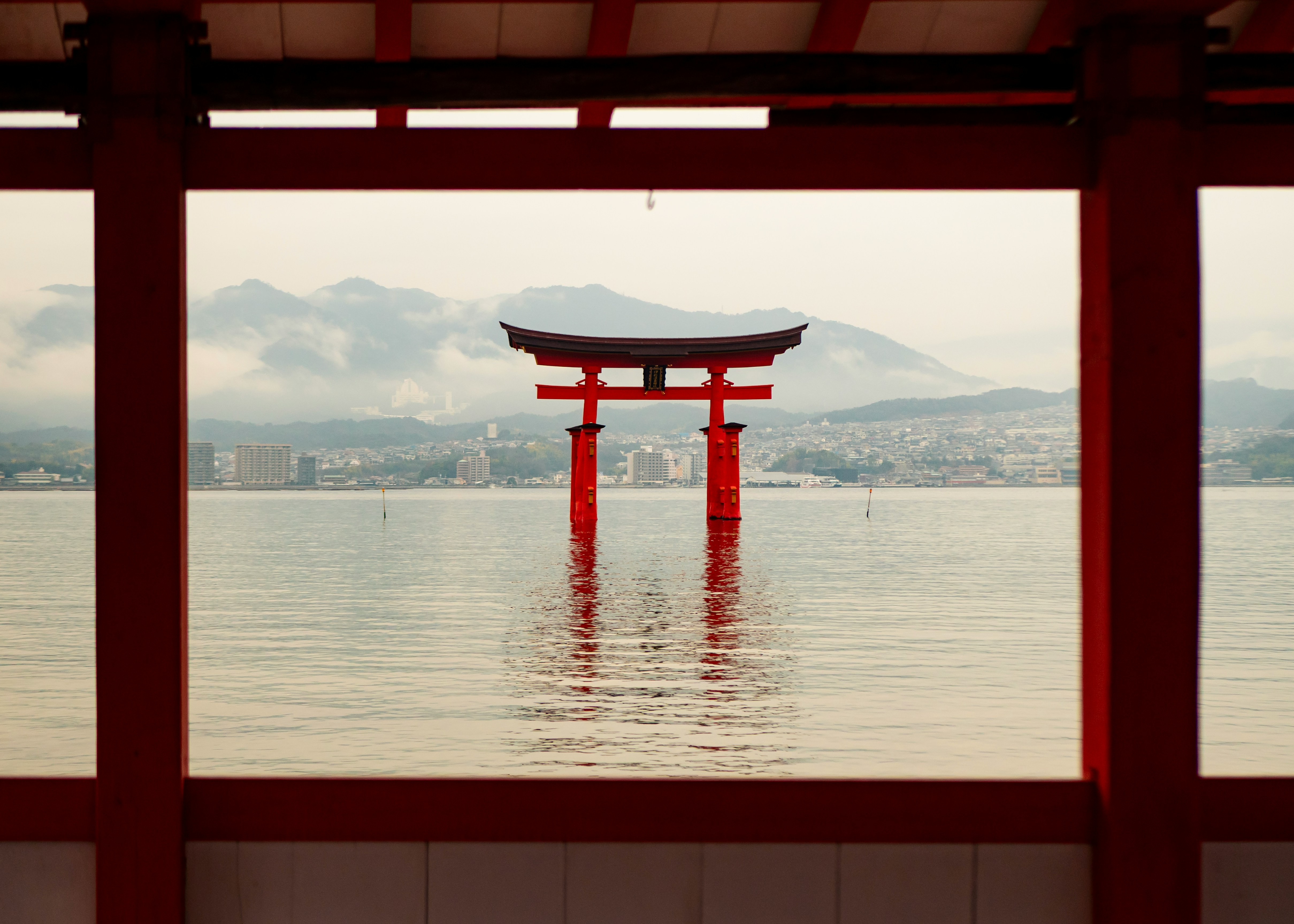 A red torii gate stands in calm water, framed by shrine columns and backed by misty mountains across the bay.