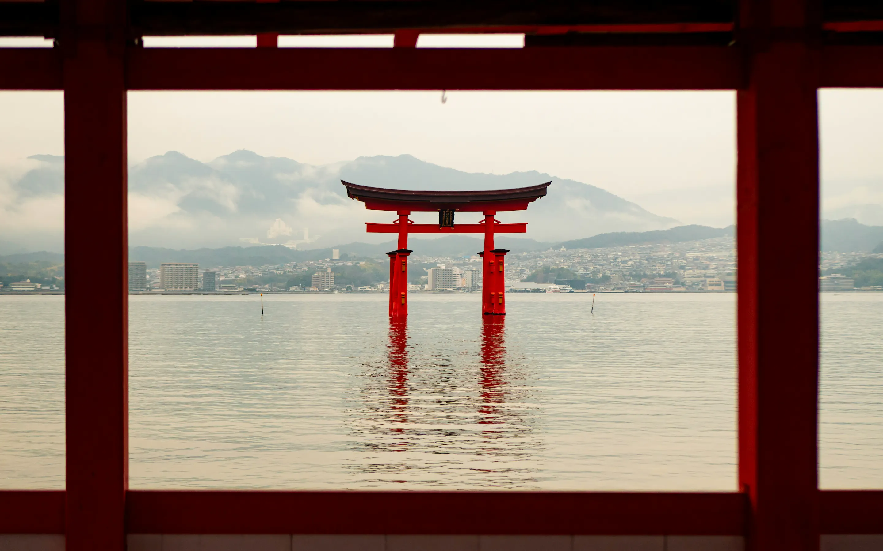 A red torii gate stands in calm water, framed by shrine columns and backed by misty mountains across the bay.