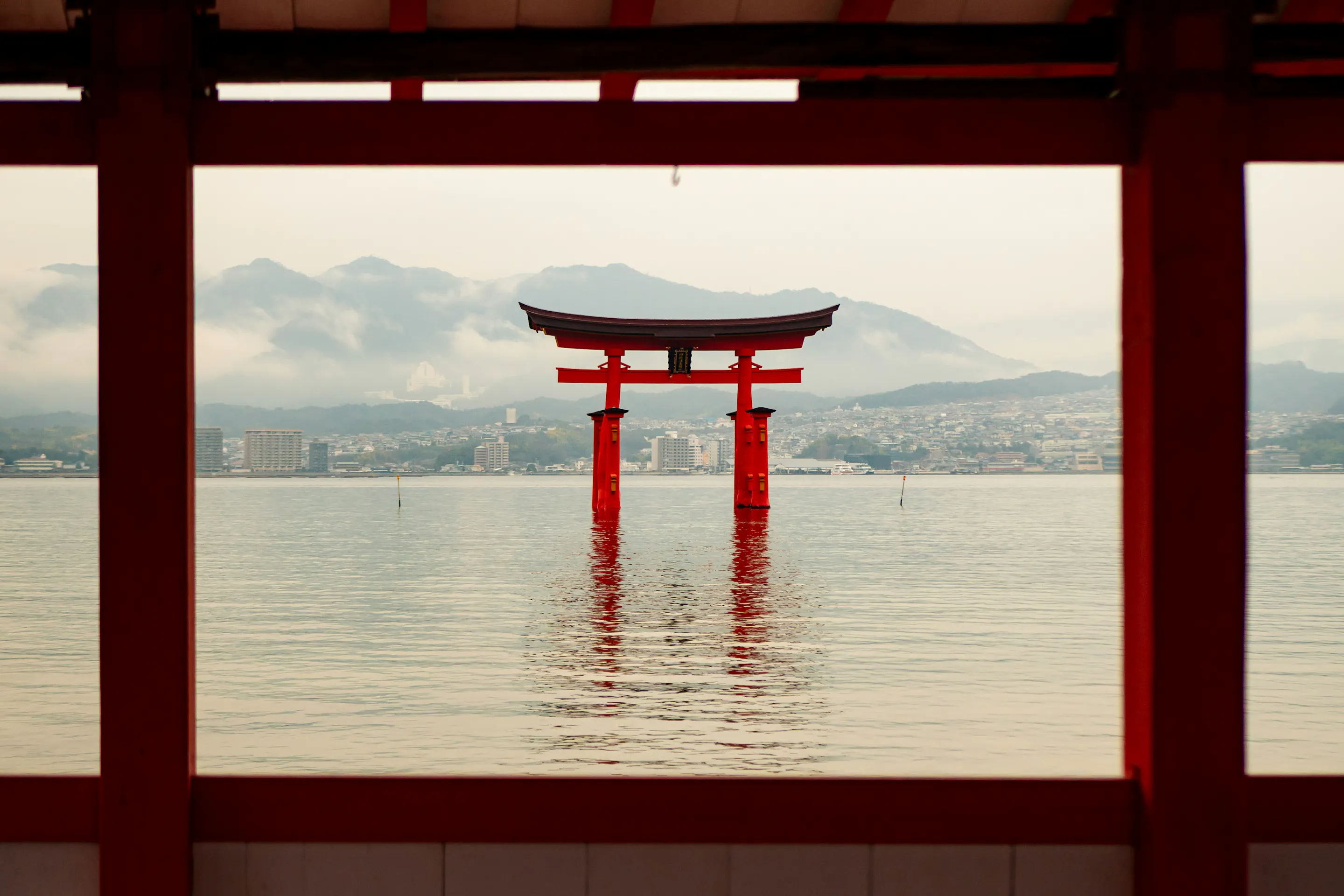 A red torii gate stands in calm water, framed by shrine columns and backed by misty mountains across the bay.