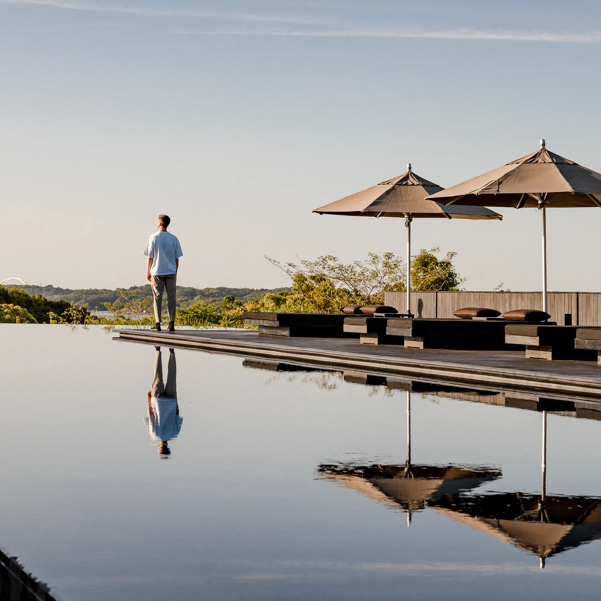 A guest stands beside Amanemu's still pool under parasols, with mirrorlike reflections and low hills beyond the deck.