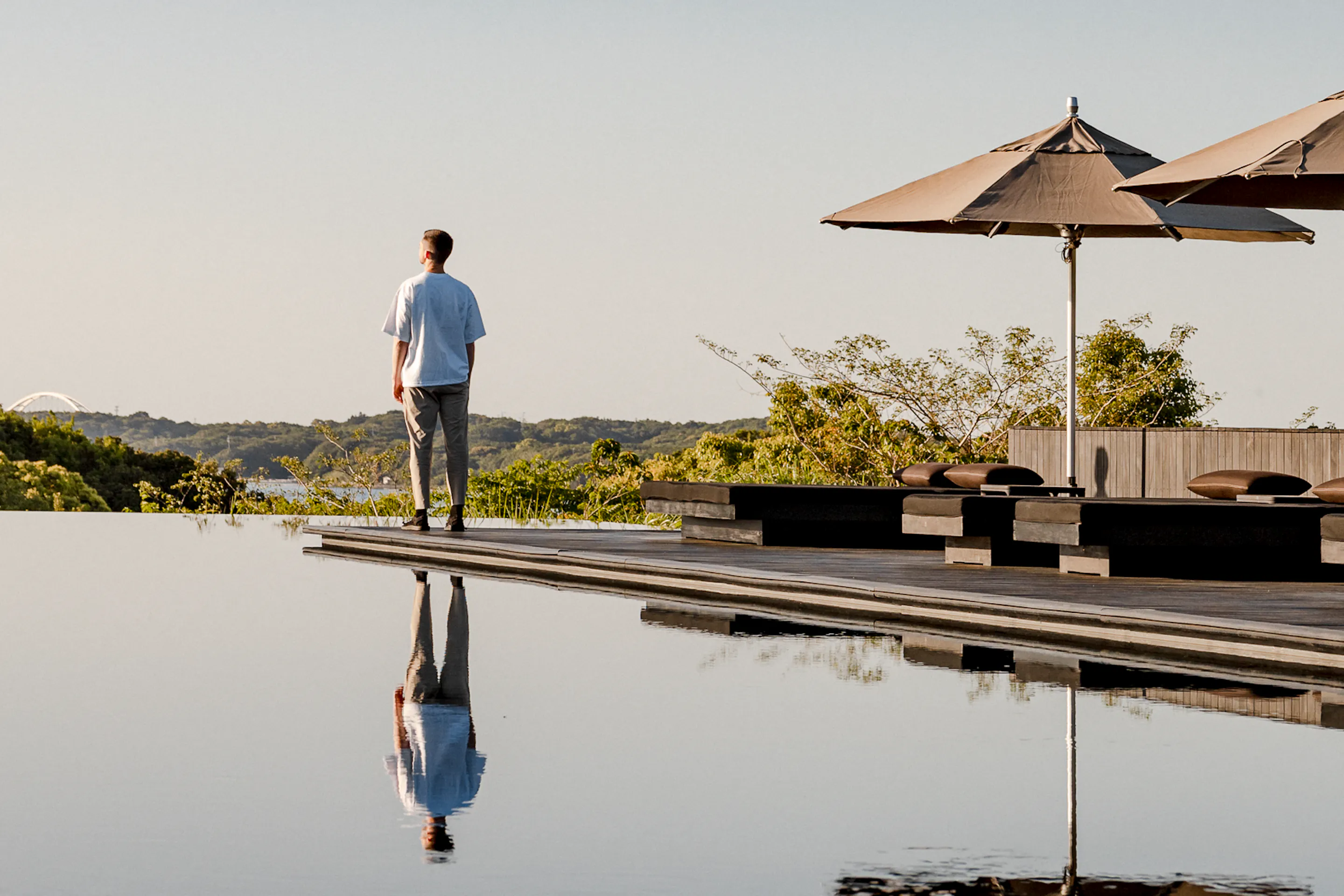 A guest stands beside Amanemu's still pool under parasols, with mirrorlike reflections and low hills beyond the deck.