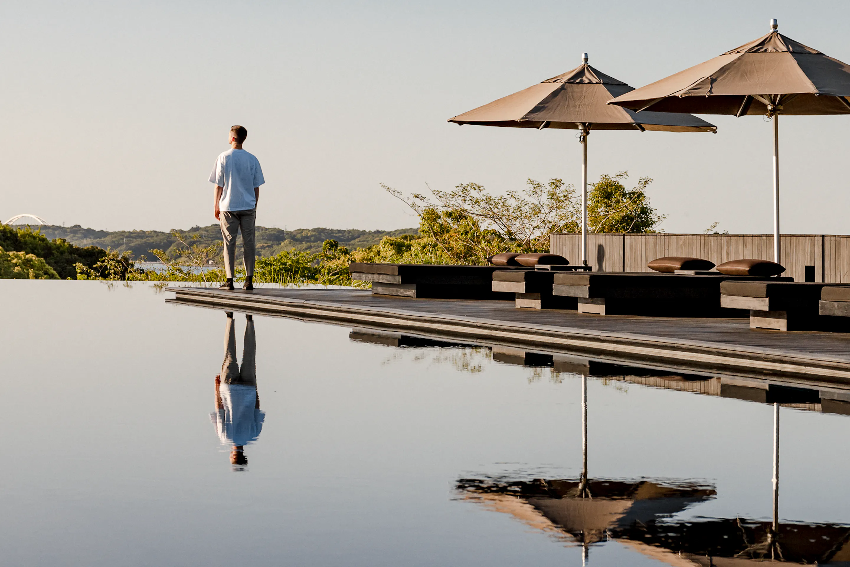 A guest stands beside Amanemu's still pool under parasols, with mirrorlike reflections and low hills beyond the deck.