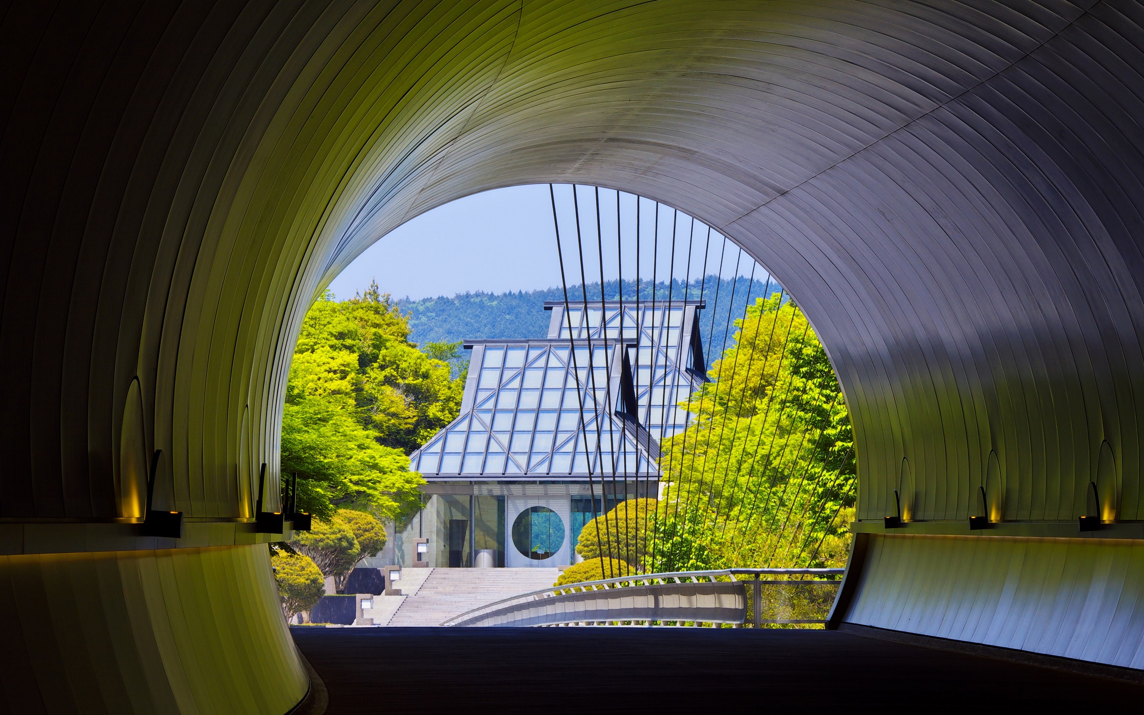 The curved tunnel at MIHO Museum frames the main building ahead, with spring trees and a bridge leading inward.