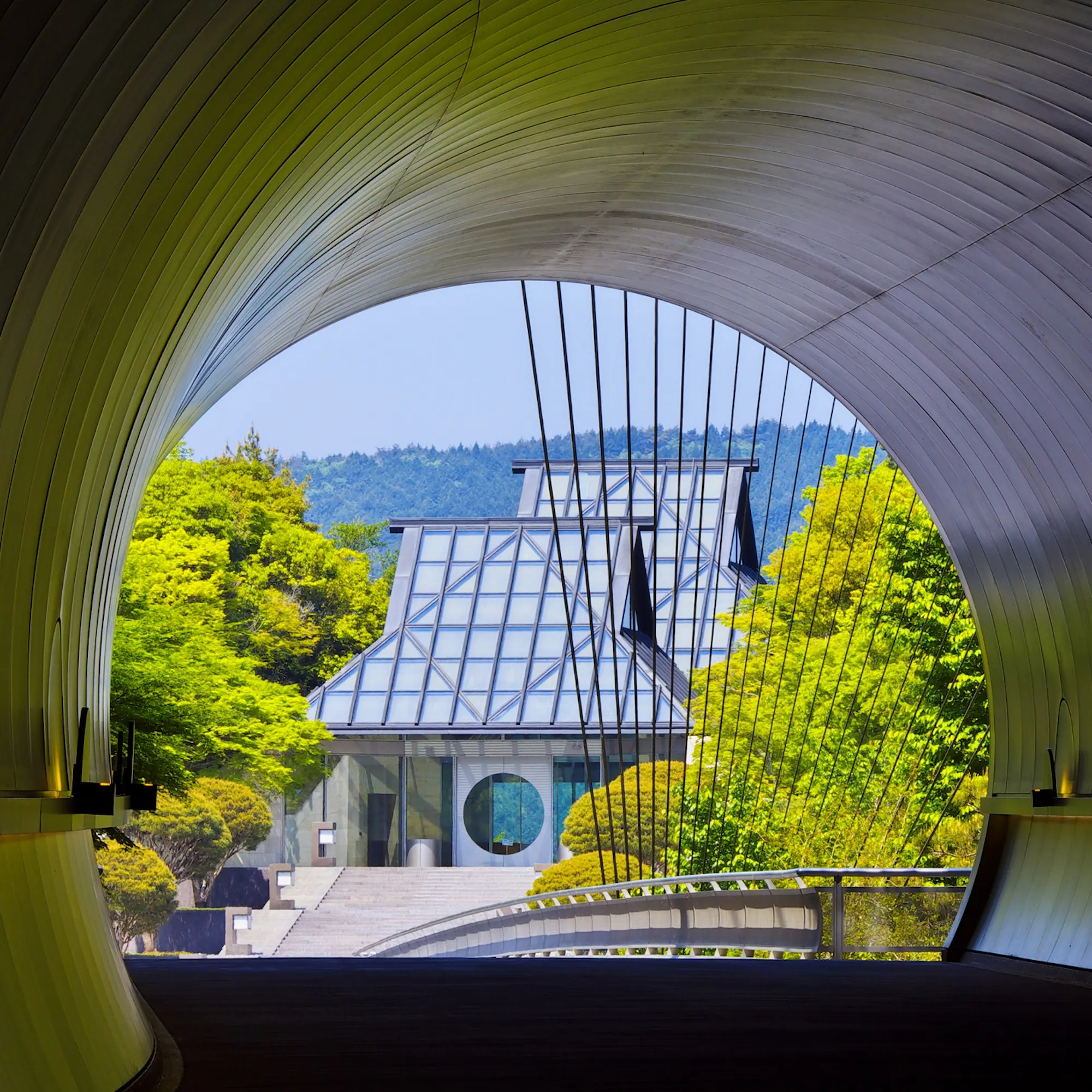 The curved tunnel at MIHO Museum frames the main building ahead, with spring trees and a bridge leading inward.