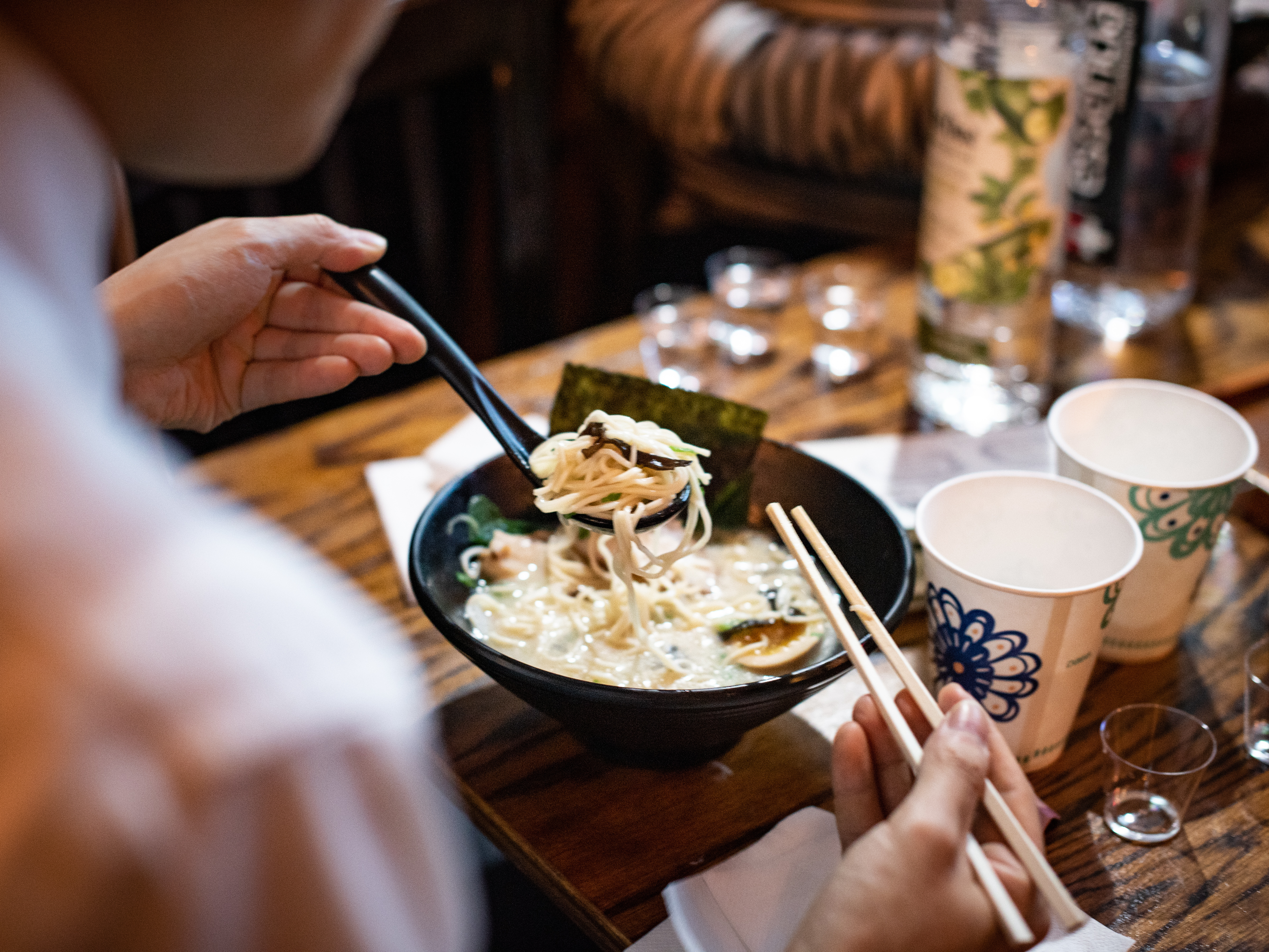 Two diners lift noodles from a steaming bowl of ramen at a wooden table set with cups, chopsticks, and drinks.