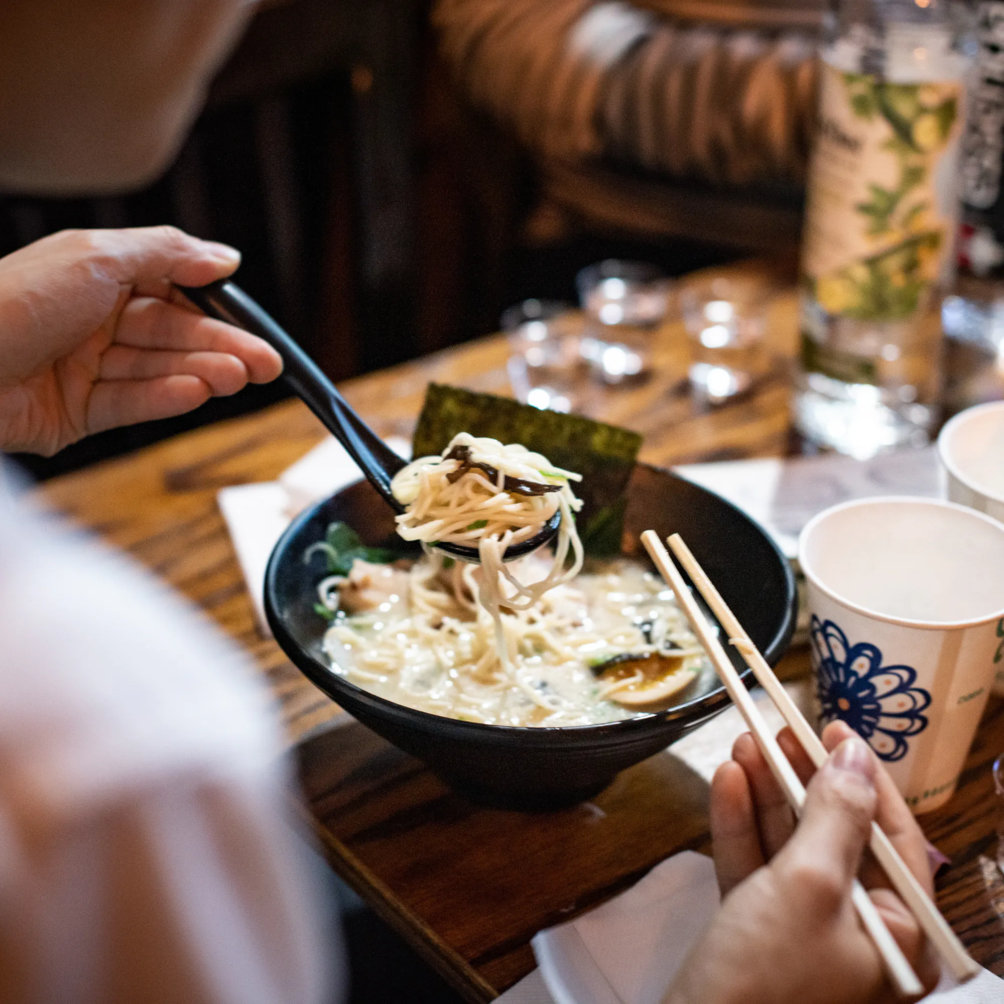 Two diners lift noodles from a steaming bowl of ramen at a wooden table set with cups, chopsticks, and drinks.